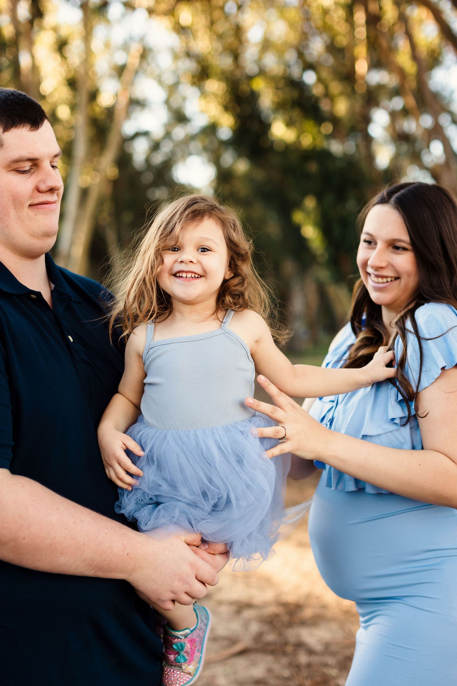 A couple in light blue and black clothing share a tender embrace outdoors during a golden sunset photoshoot.
