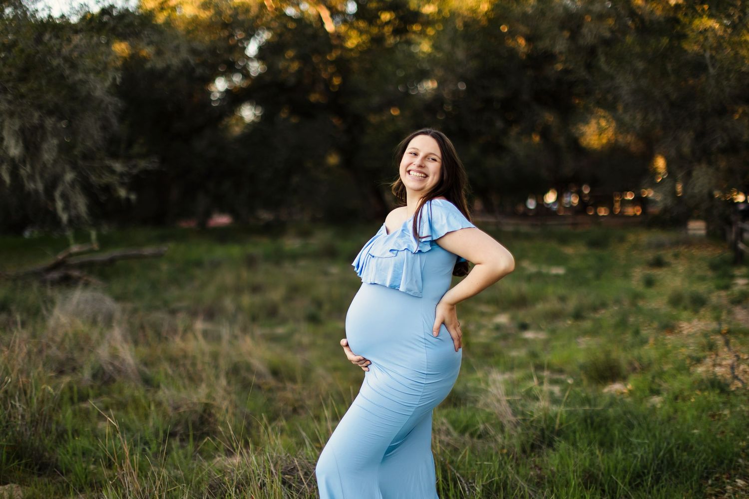 Person wearing a light blue dress poses outdoors at sunset in a natural grassy setting.