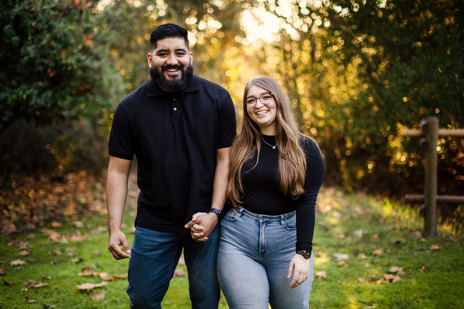 A couple stands together smiling on a grass lawn during golden hour with autumn trees in the background.