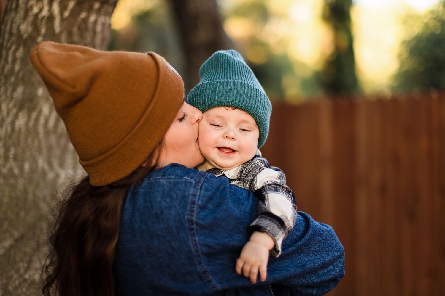 A smiling baby in a teal beanie is held by someone wearing a navy blue sweater during a sunny outdoor moment.
