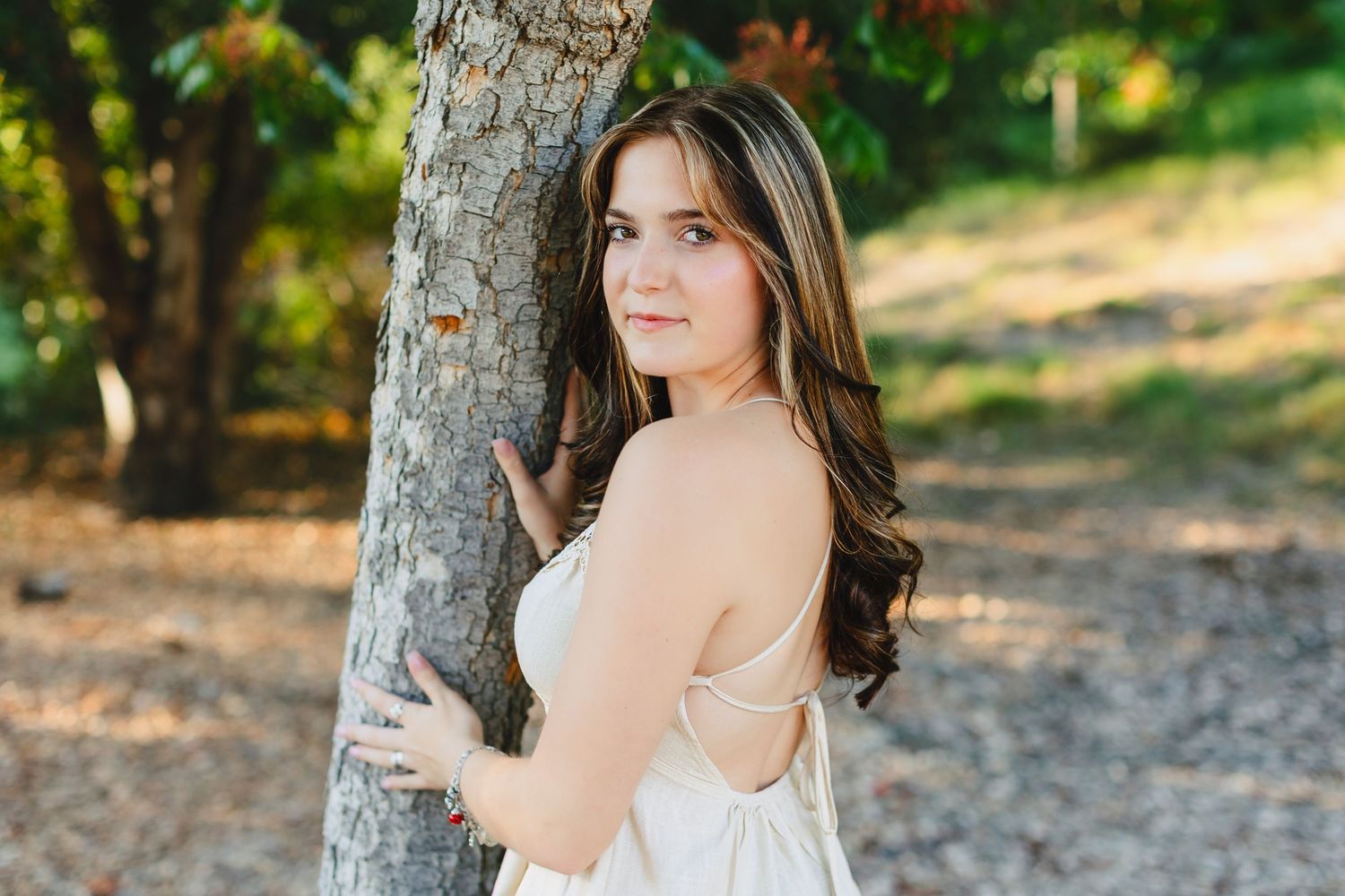 A person in a white dress poses against a tree trunk in a sunlit outdoor setting during golden hour.