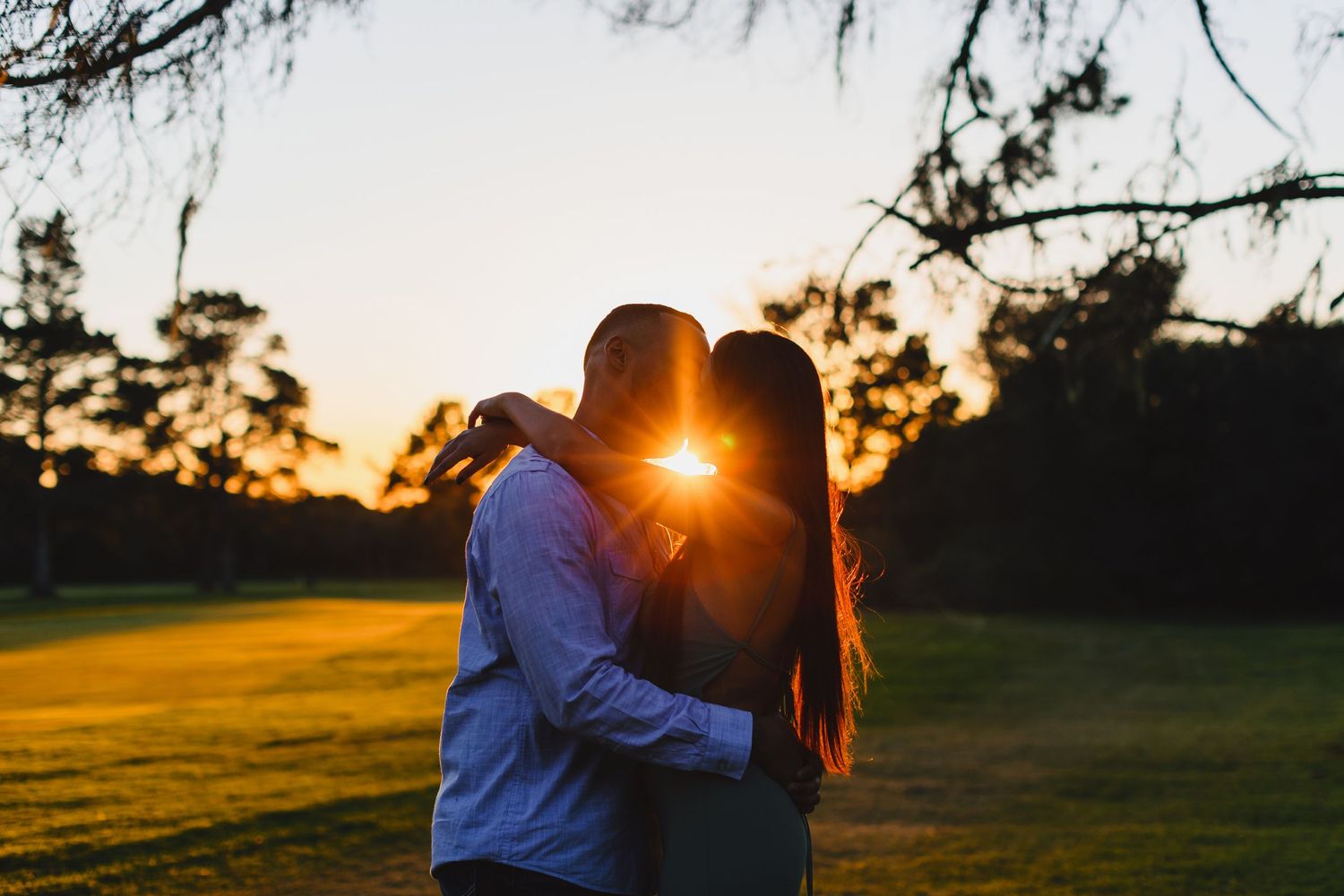 Silhouetted couple embracing in a field during golden hour with sun flare between them.