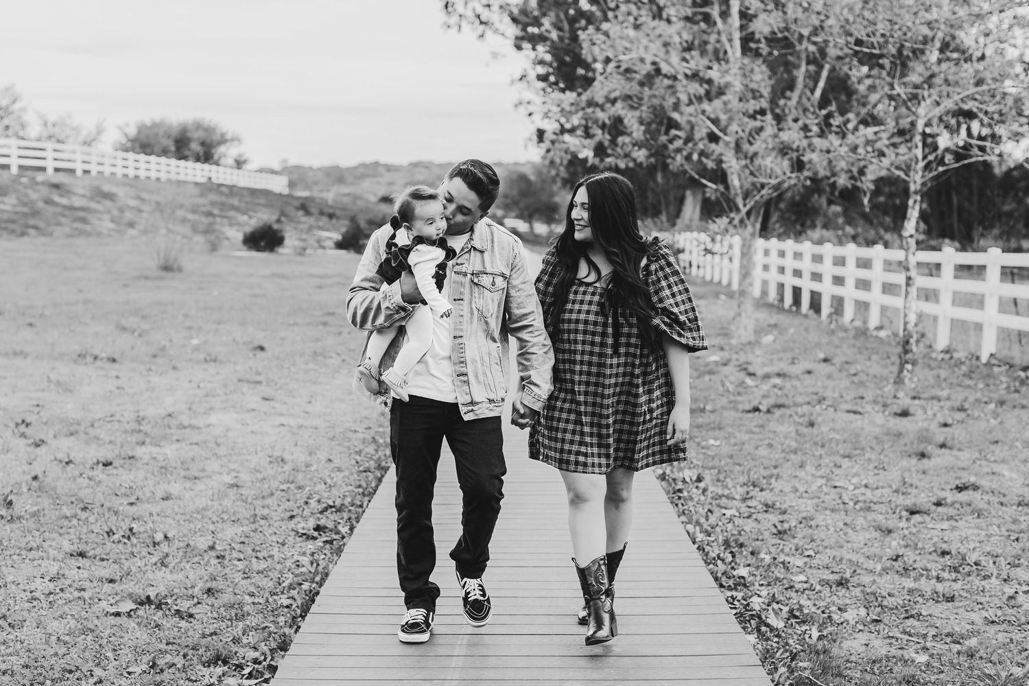 Black and white photo of a family walking on a wooden path near a white fence in a rural setting.