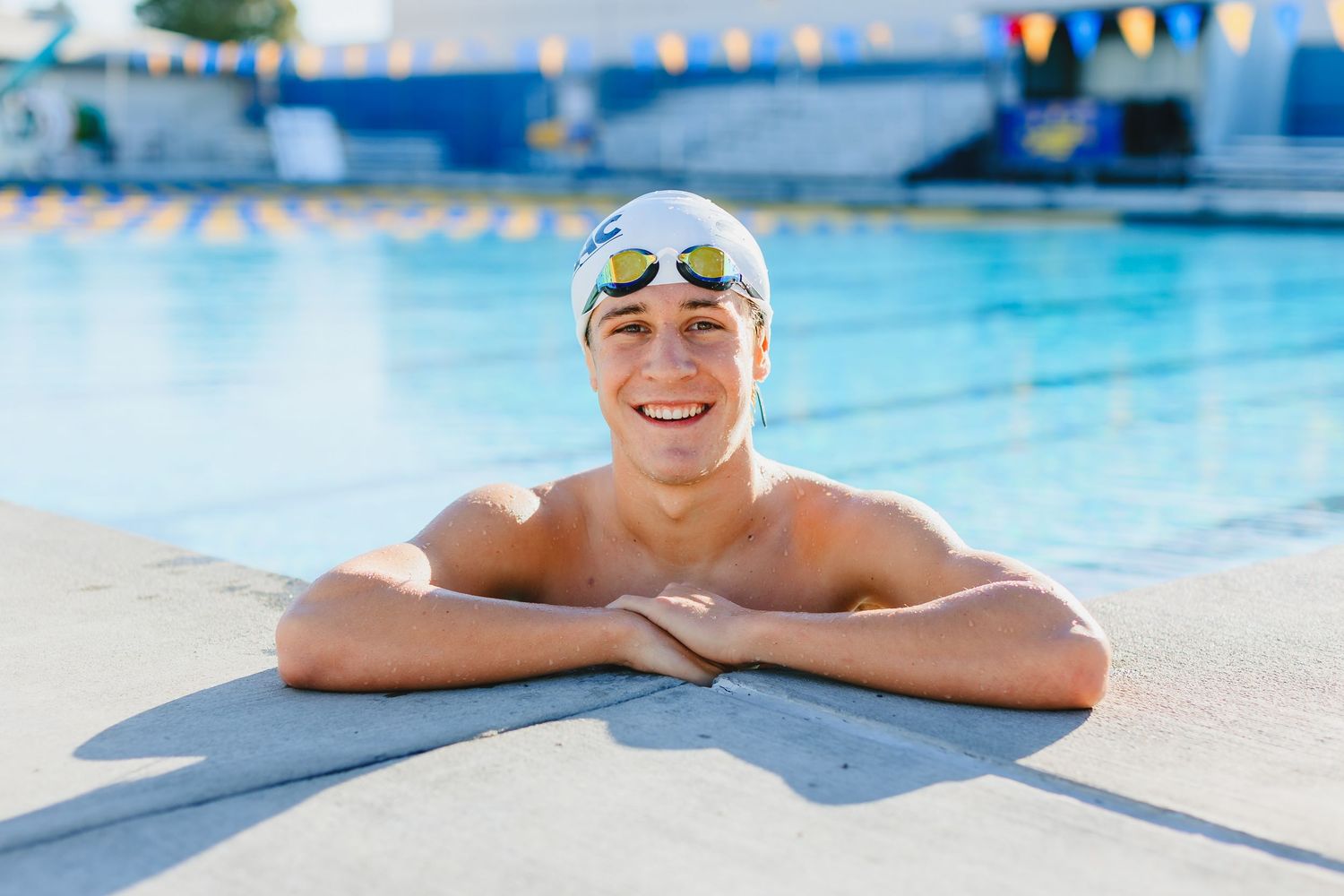 Smiling teenage male swimmer leaning on pool edge wearing white swim cap and goggles at an outdoor swimming pool.