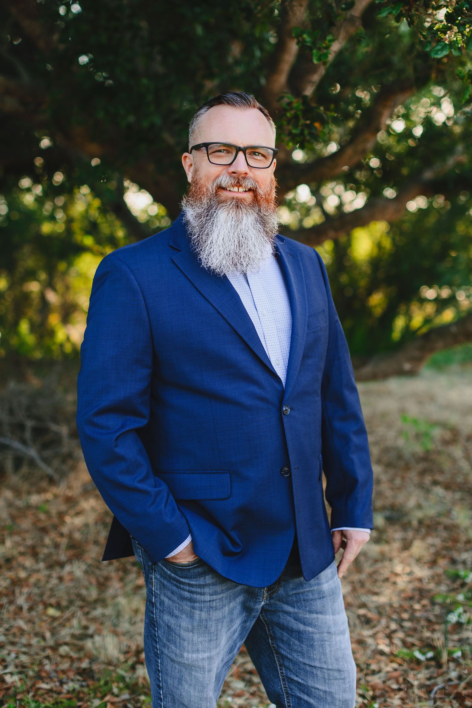 Professional in navy blazer and glasses poses against green foliage background.