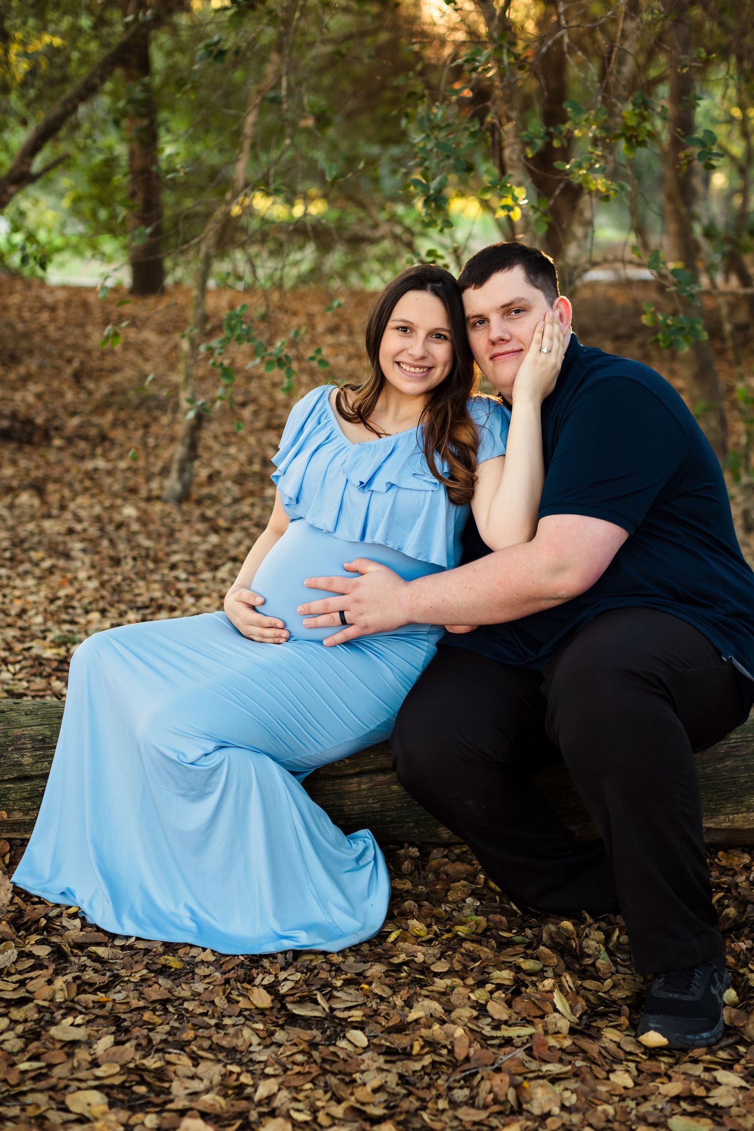 Couple in autumn setting during maternity photoshoot with person wearing light blue dress sitting on fallen leaves.