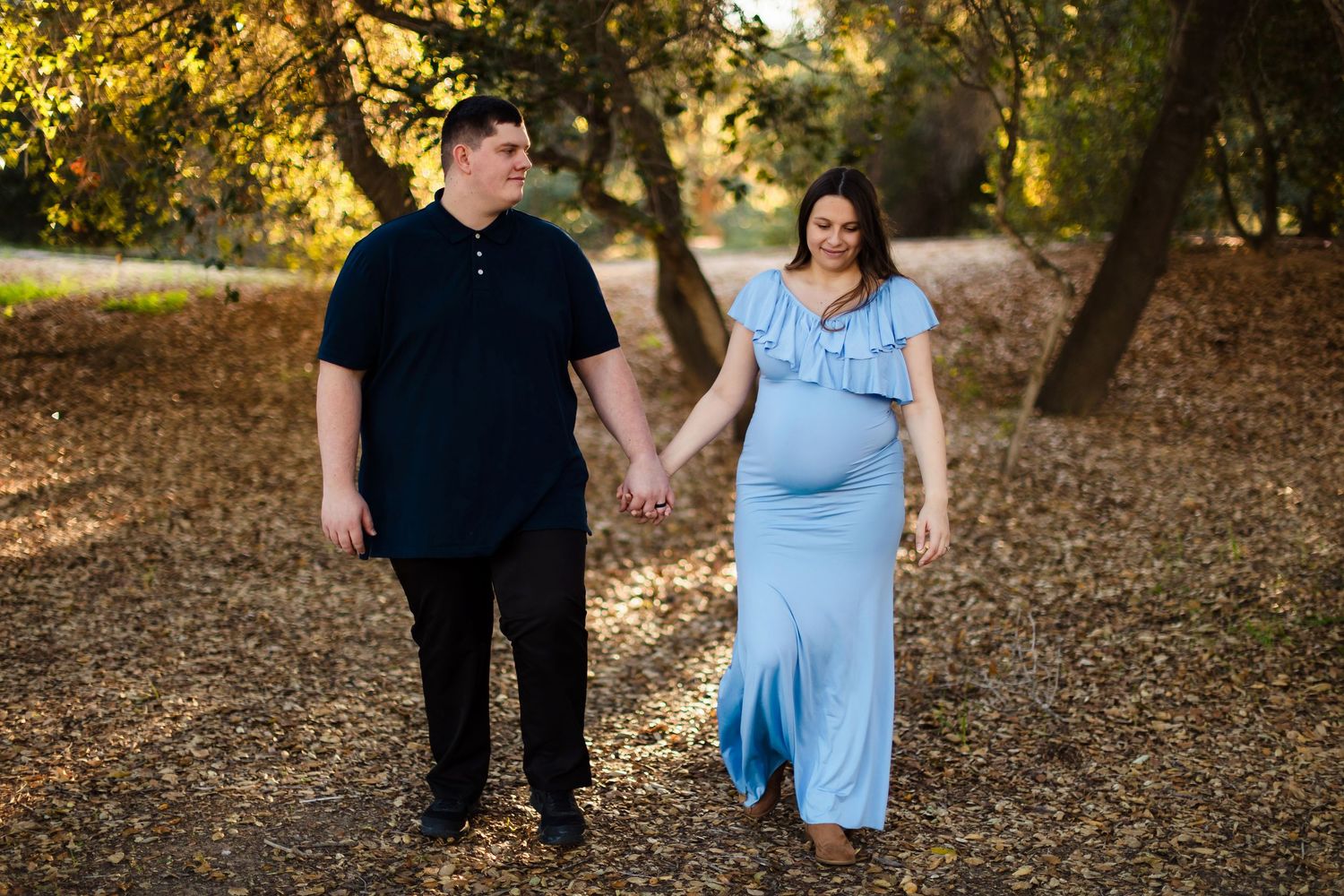 A couple enjoying a romantic autumn walk through a sun-dappled wooded path with fallen leaves around them.