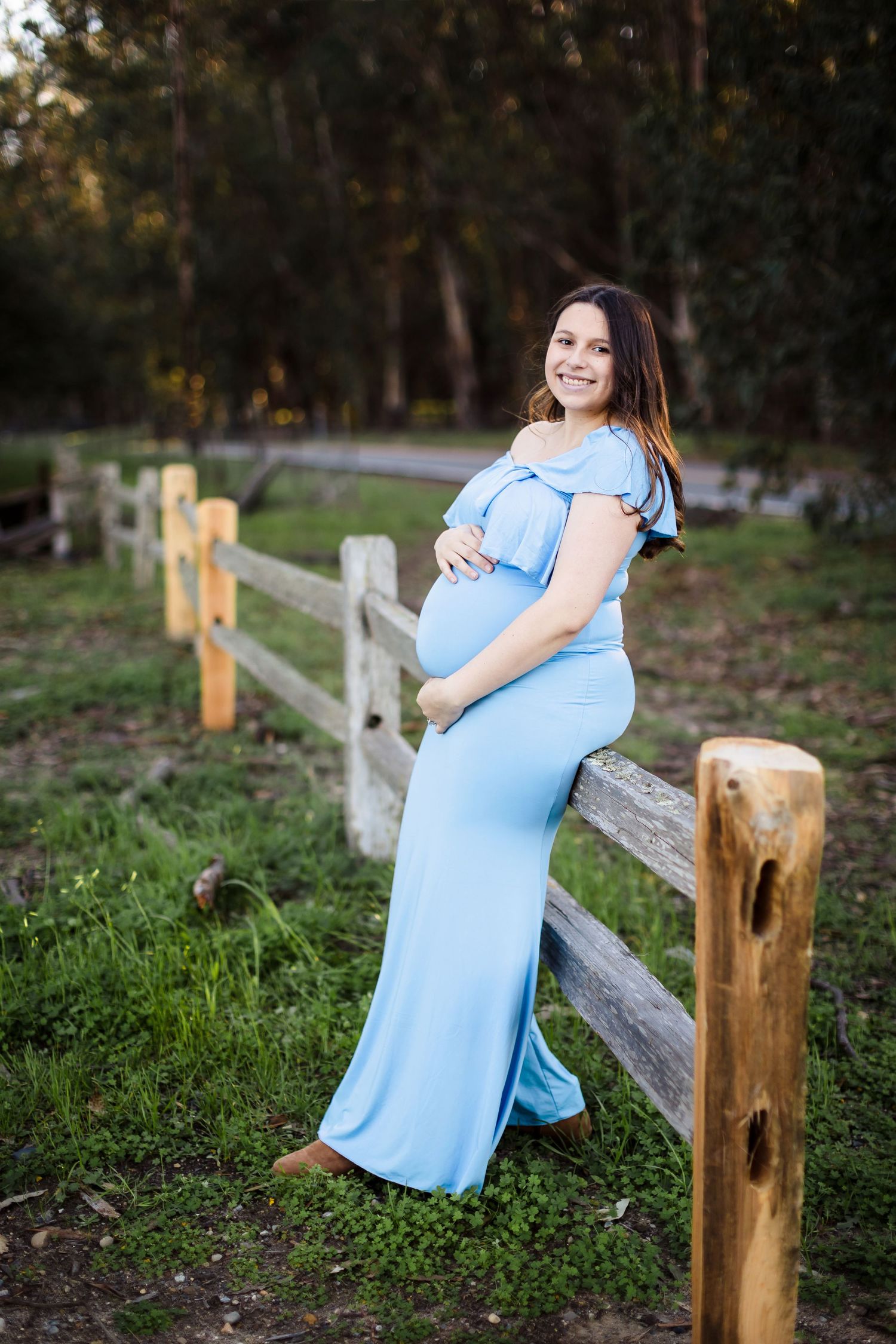 A light blue maternity gown is showcased against a rustic wooden fence in an outdoor natural setting at sunset.