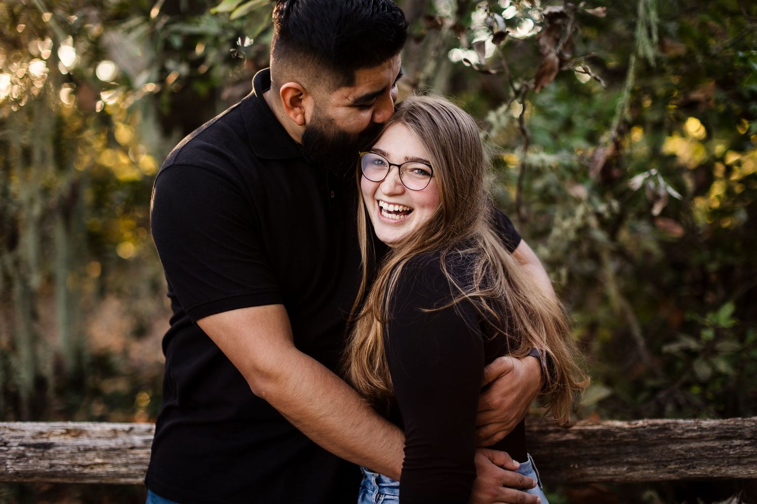A couple shares playful and romantic moments together on a wooden bench surrounded by trees and autumn foliage.