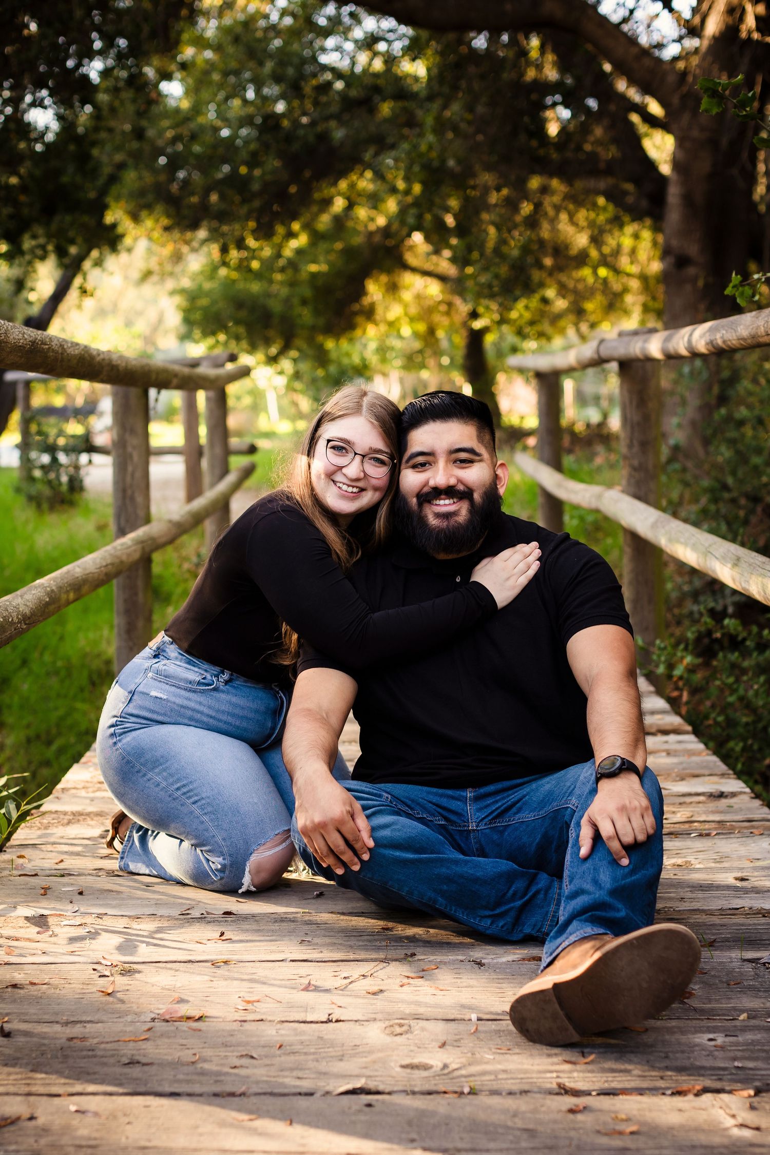 A couple relaxes and poses together on a wooden boardwalk surrounded by trees during a casual outdoor photoshoot.