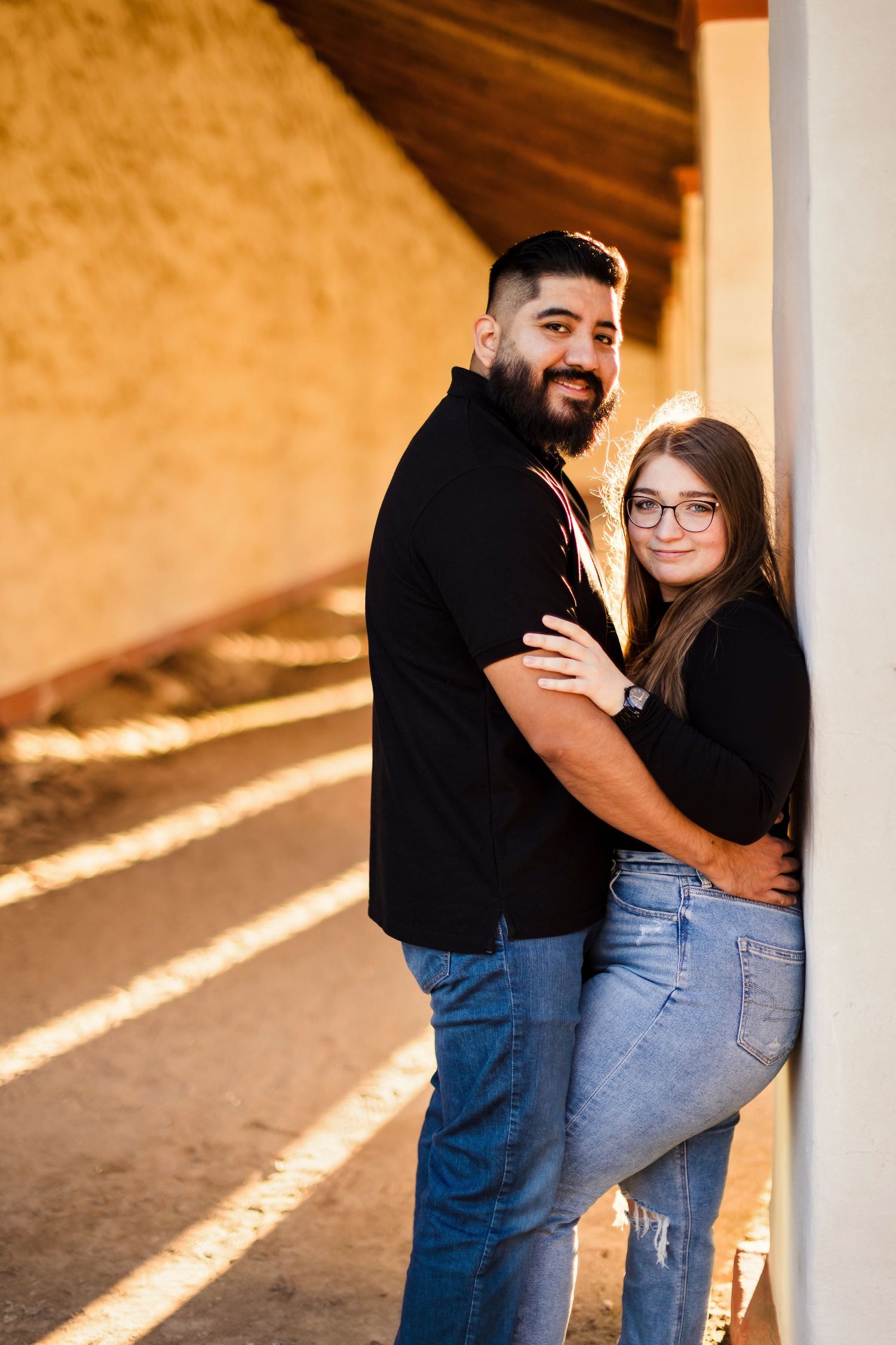 A couple embraces lovingly while standing in a sunlit concrete corridor with columns.