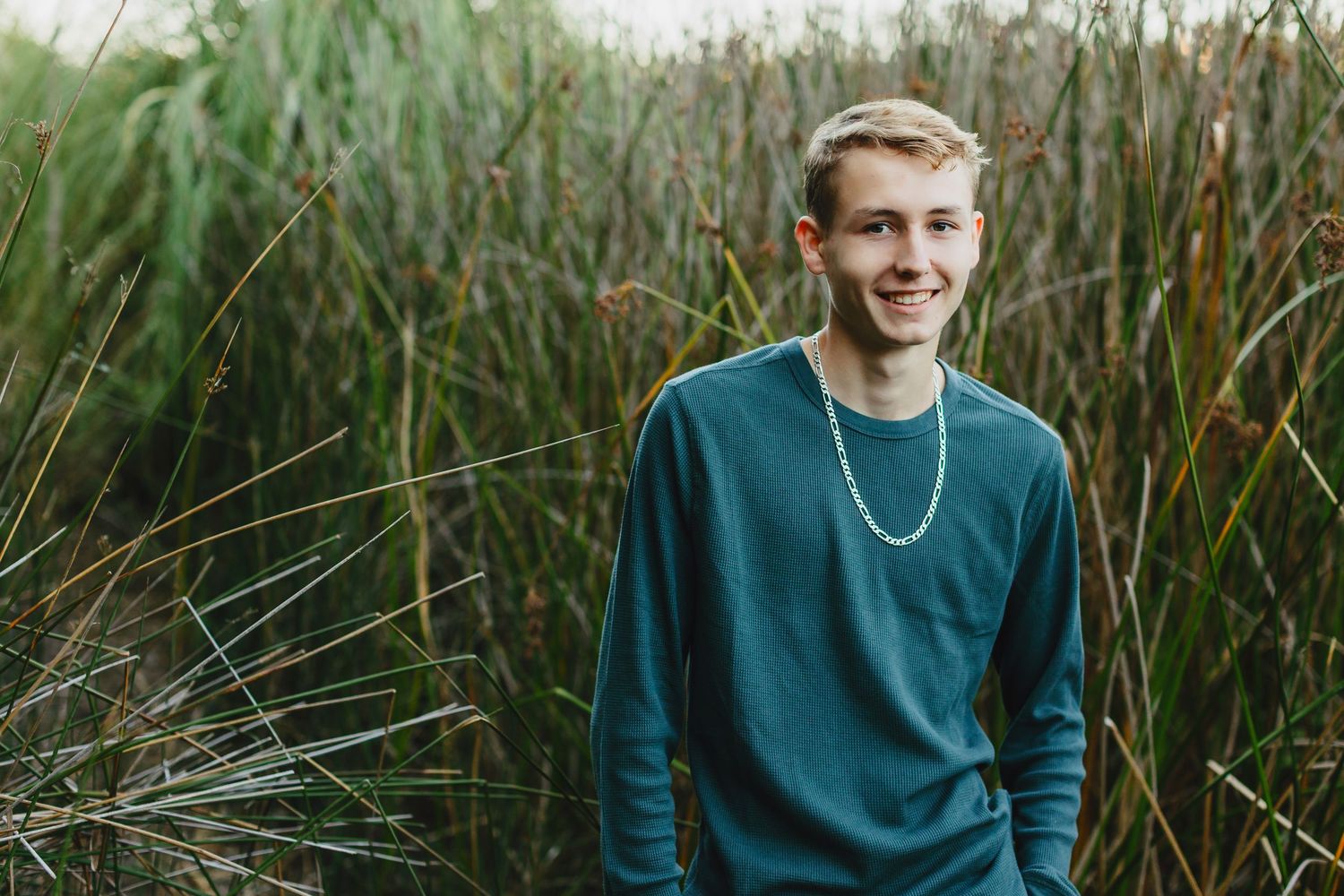 A person in a teal sweater smiles while sitting among tall grass and reeds in an outdoor nature setting.