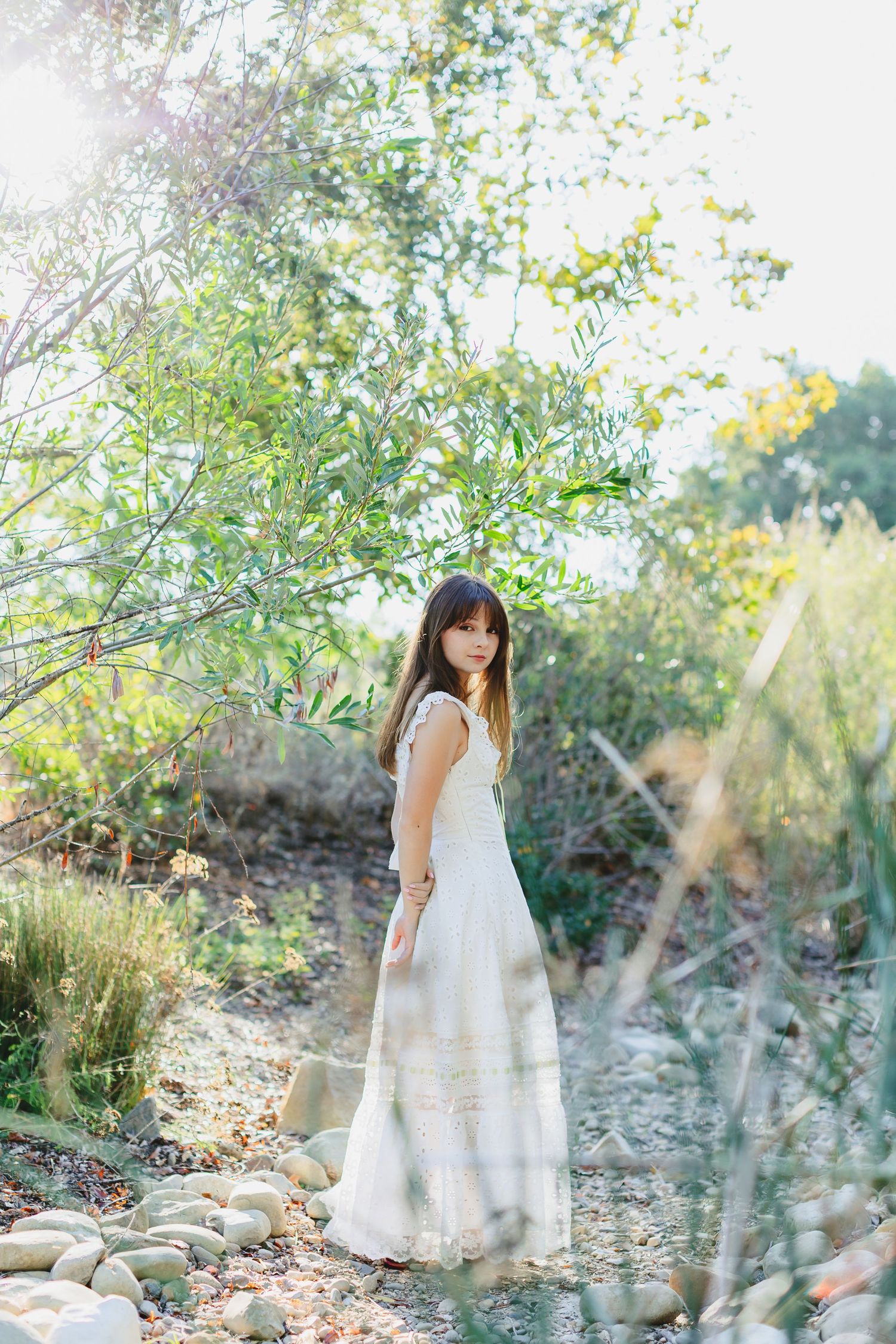 Young woman in white dress standing on rocky path surrounded by lush green trees in sunlit garden.
