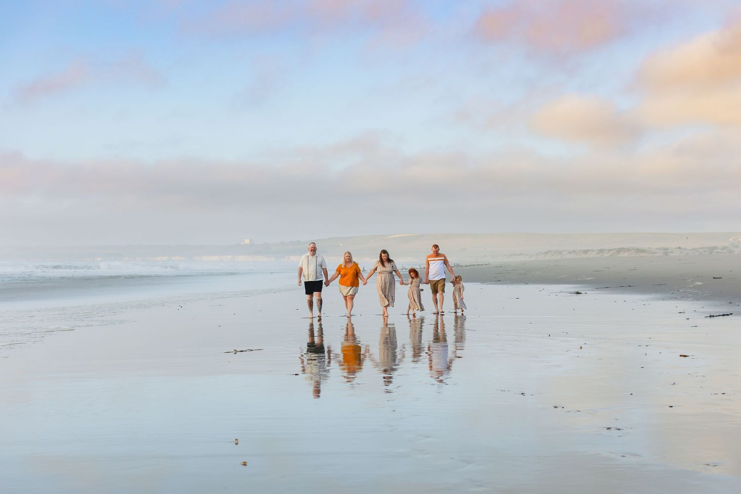 Group of people walking along a misty beach at sunset, their reflections mirrored in the wet sand.