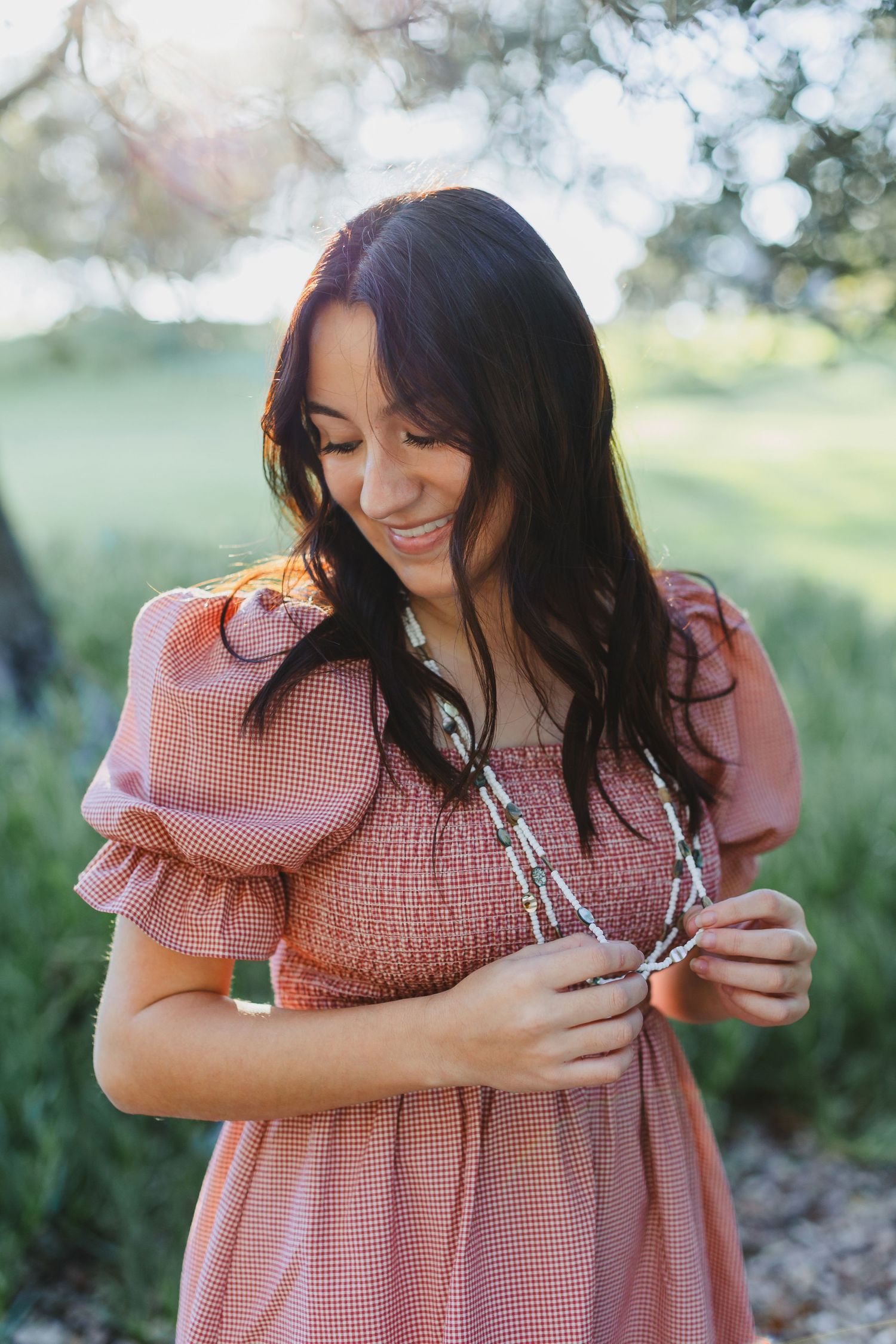 Person in pink gingham dress smiling while holding necklace outdoors with bokeh background.