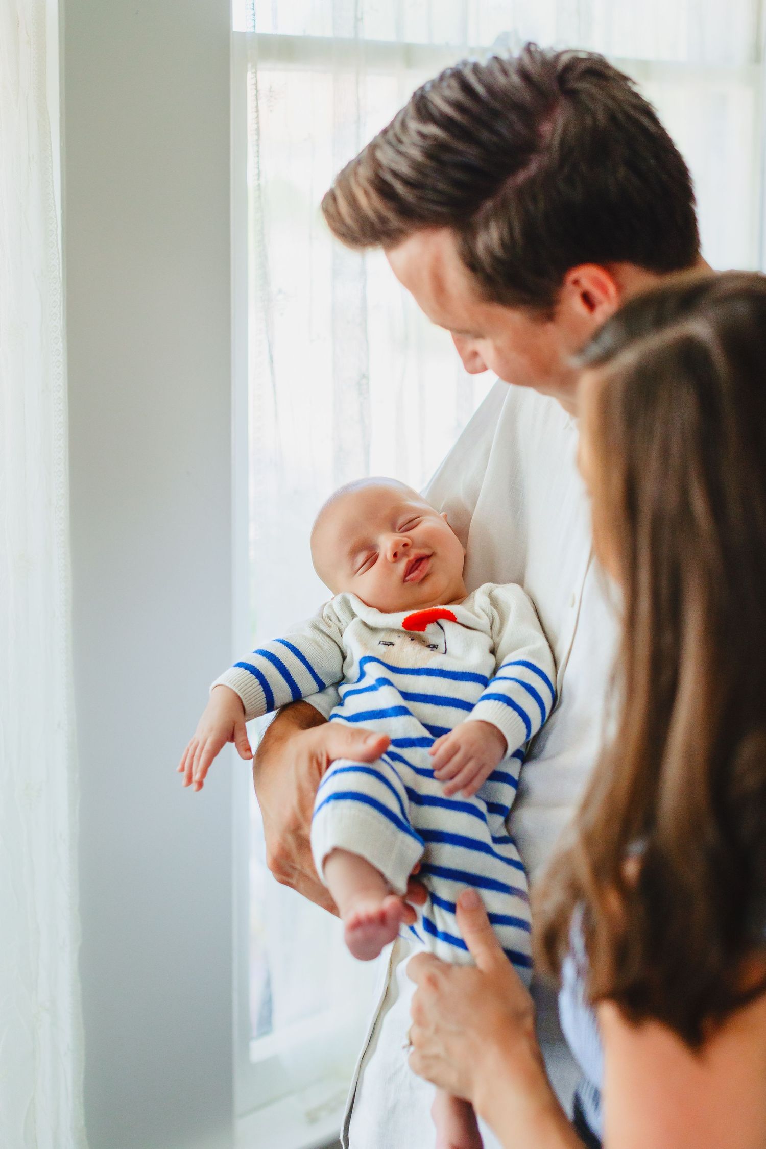 Parents admiring their smiling newborn baby dressed in blue striped onesie near a bright window.