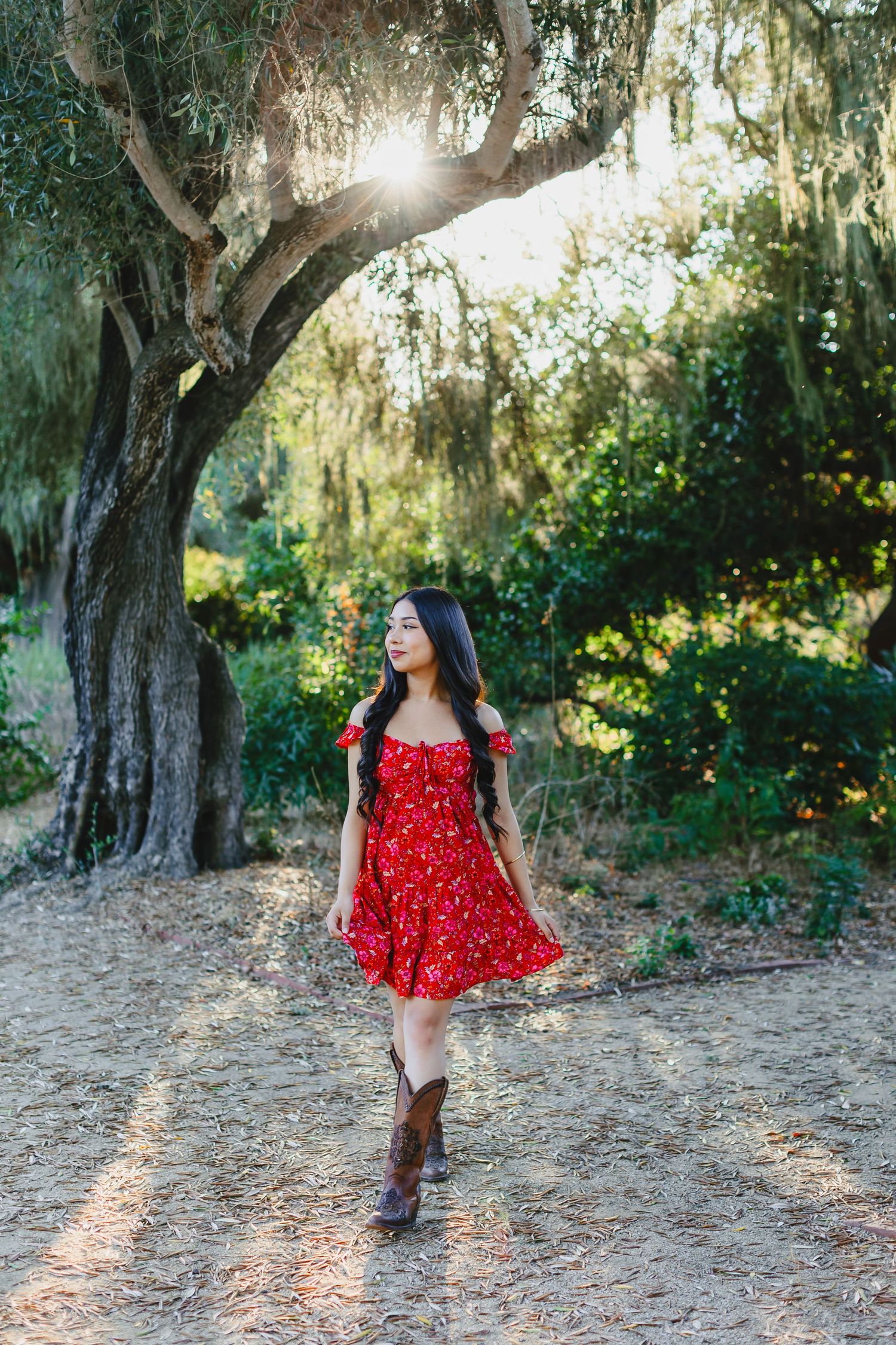 Young woman in red floral dress and cowboy boots walking under sunlit oak tree on dirt path.