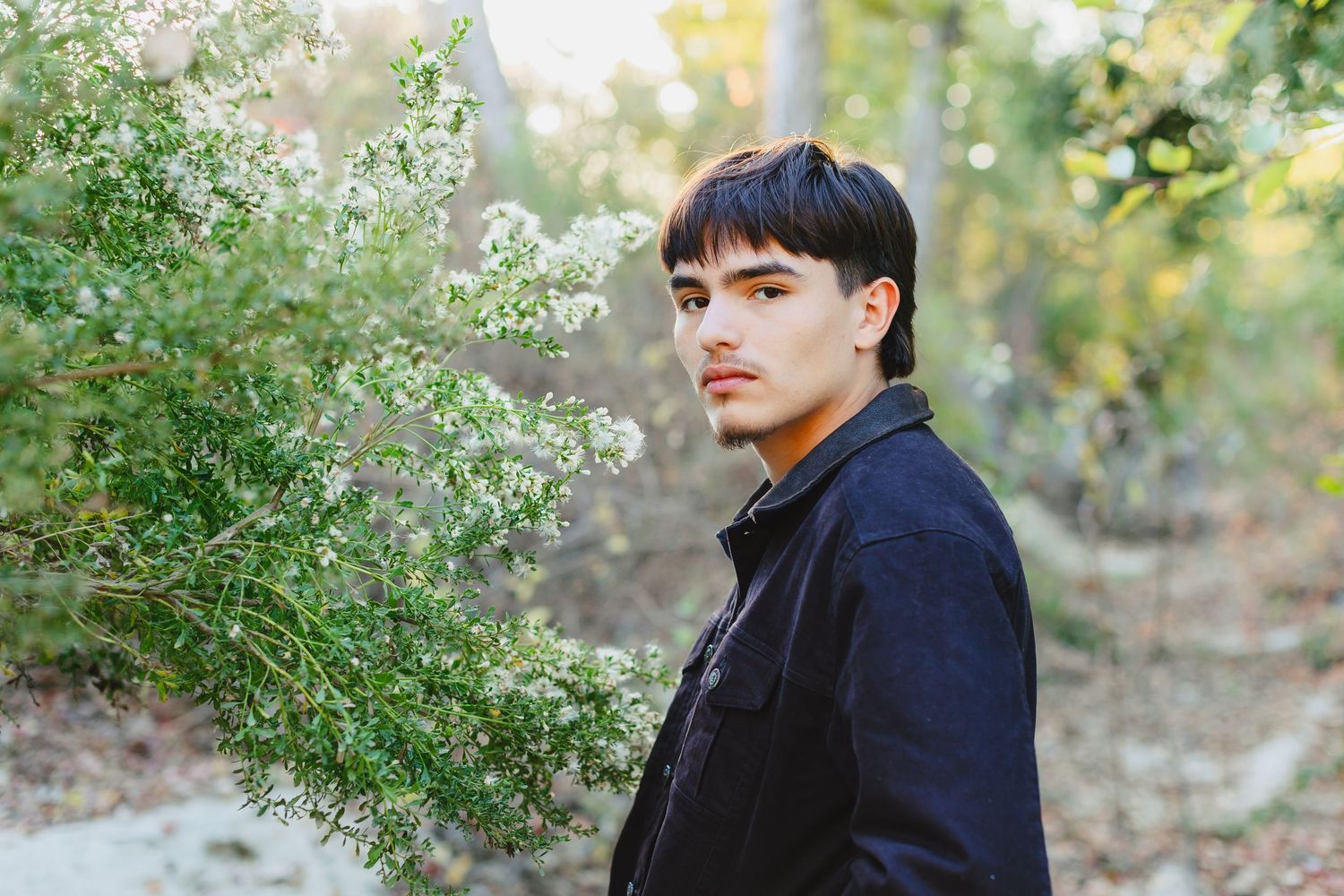 Young man with dark hair in black jacket stands beside blooming white shrubs in natural setting.