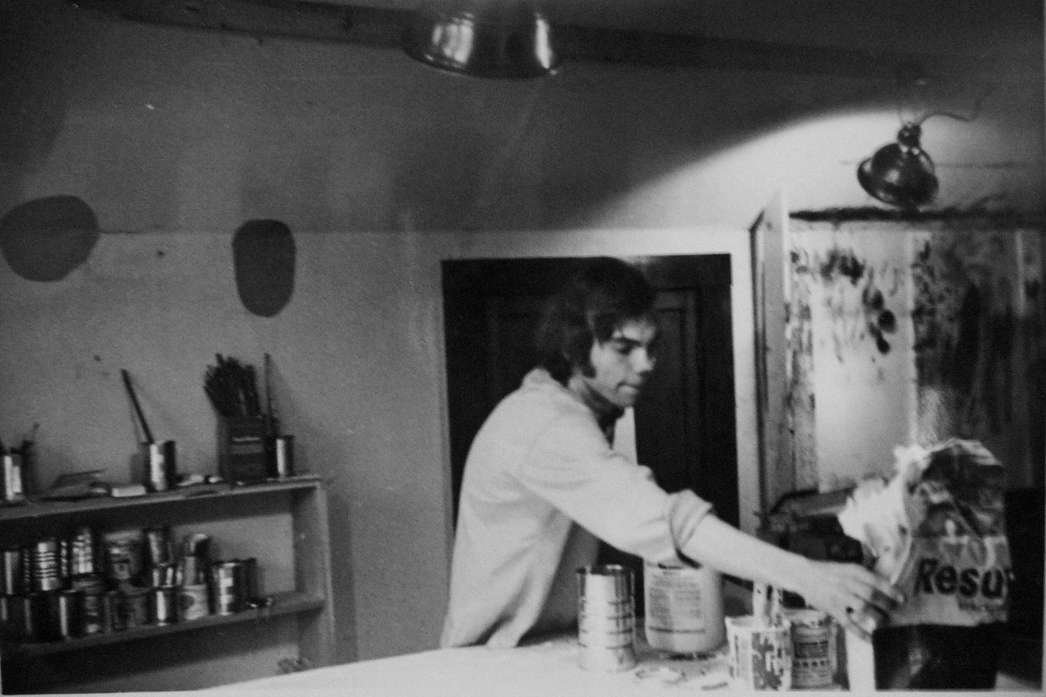A vintage black and white photograph shows someone working behind a kitchen counter with shelves of canisters visible.