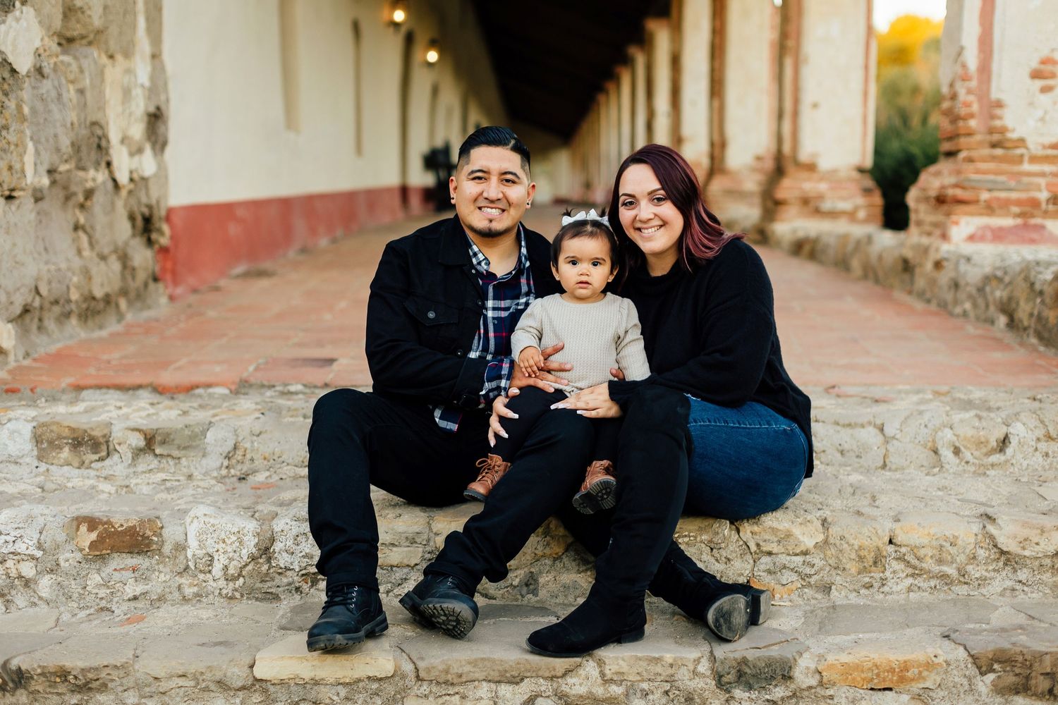 Family of three sitting together on stone steps along a rustic colonial style walkway.