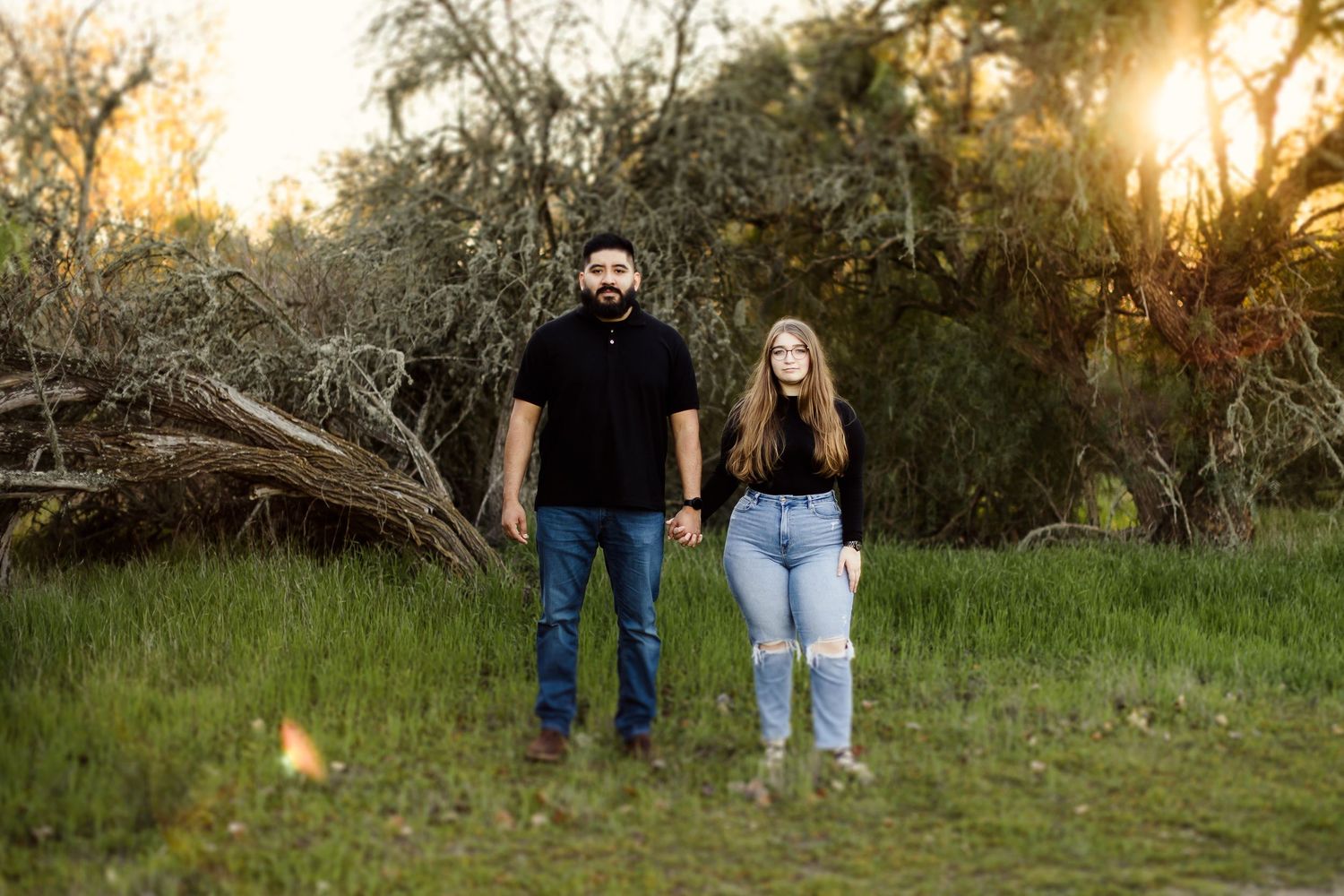 A couple poses together in warm sunlit meadow during golden hour with tall grass and trees in the background.