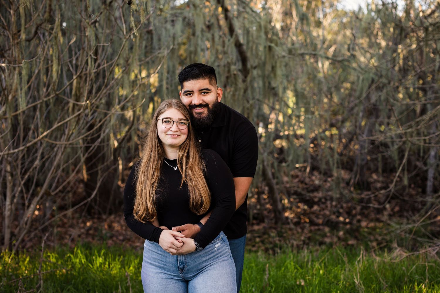 Two people pose together outdoors in a sunlit wooded area against a natural backdrop.