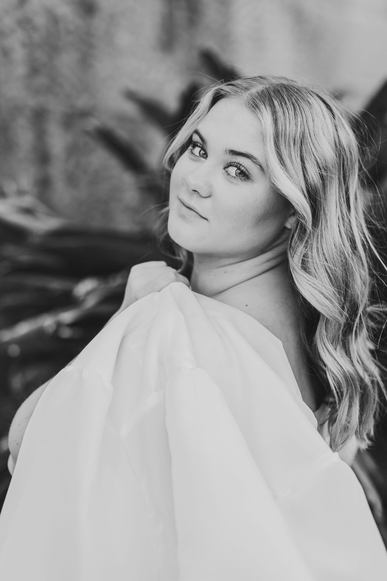 Black and white portrait of blonde woman in white off-shoulder top smiling over her shoulder.