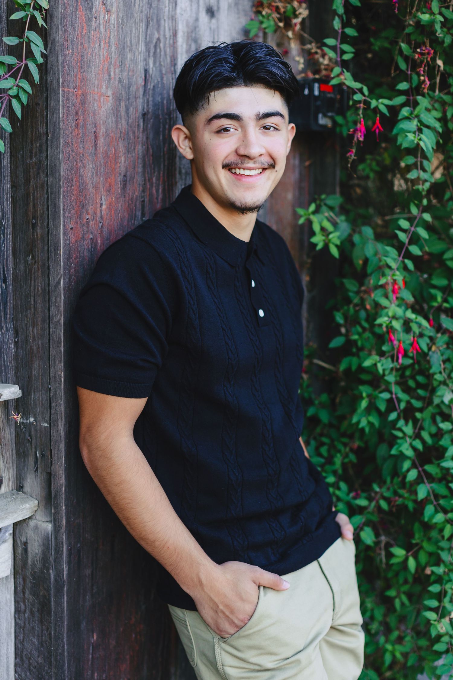 Smiling young man in black polo shirt leans against wooden wall with flowering vines.