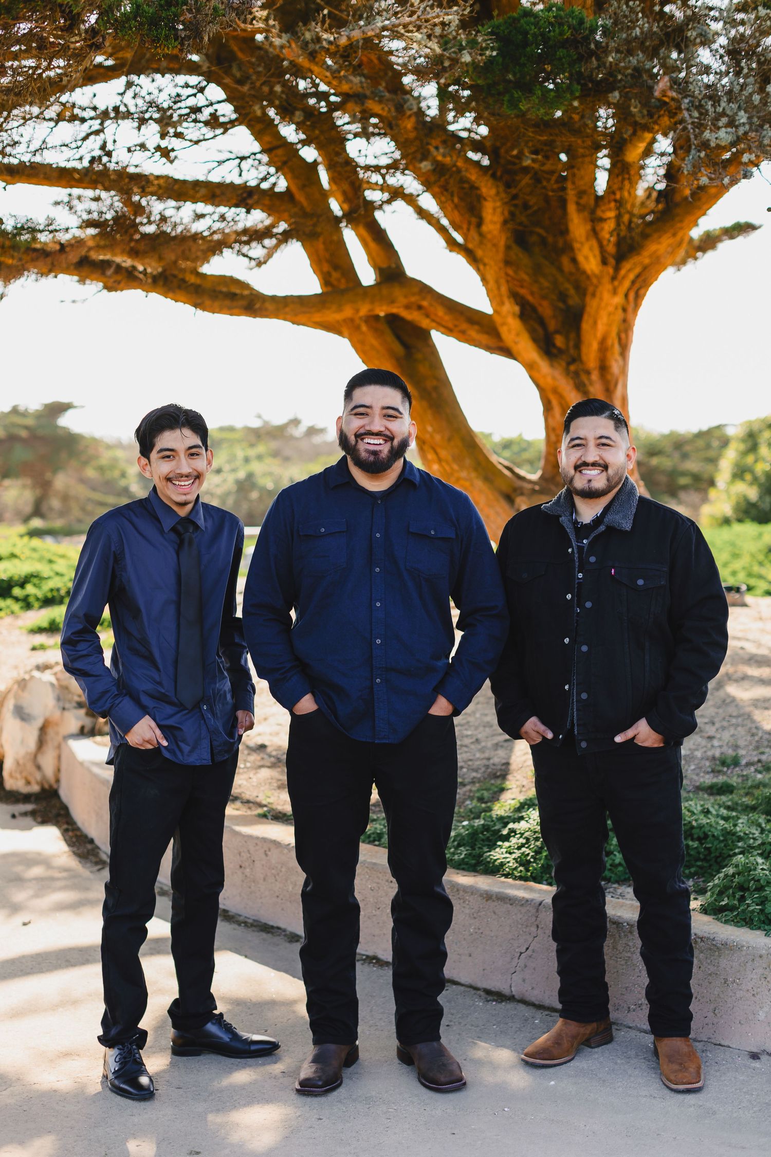 Three men in dark casual attire pose together under a large tree.