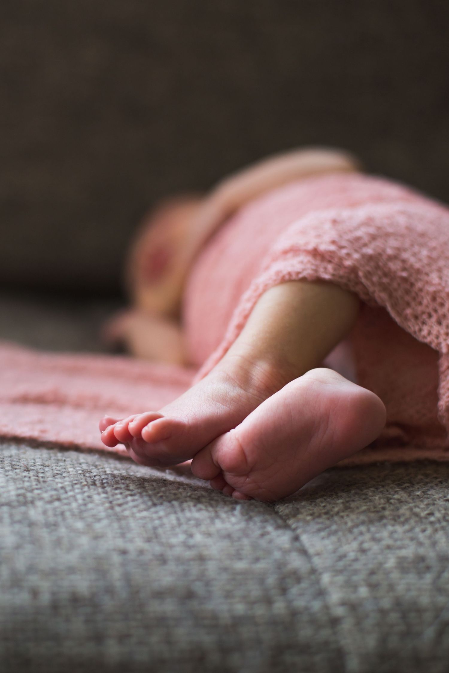 Small delicate baby feet peek out from under a pink blanket on a gray couch.