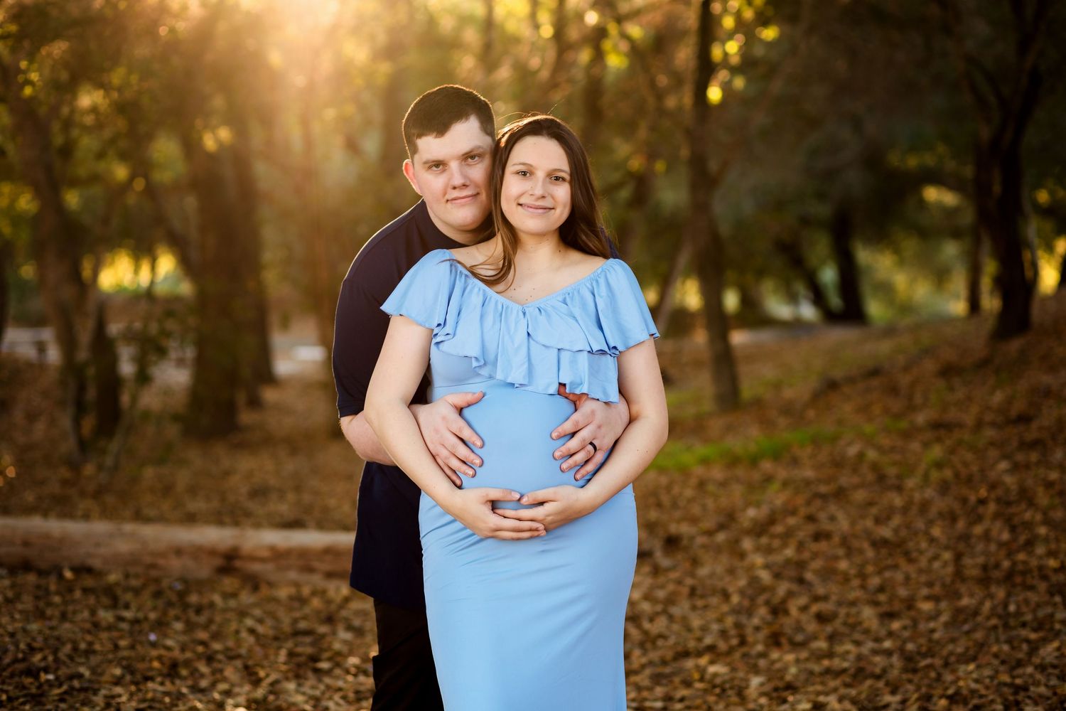 A maternity photography session captured in a wooded park during golden hour with sunlight filtering through autumn leaves.