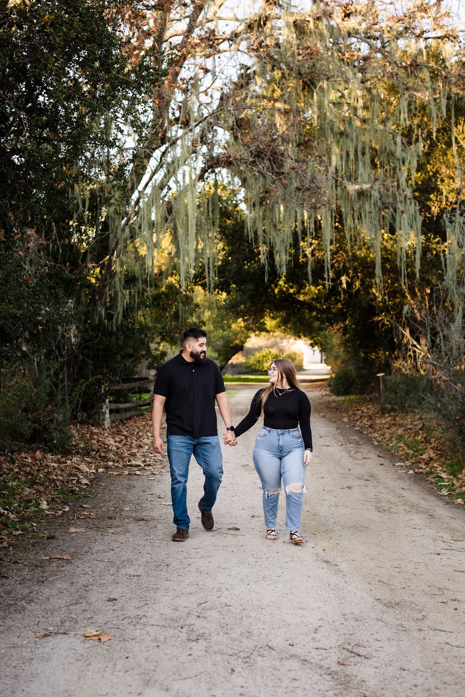 Two people walk together holding hands on a dirt path surrounded by trees with Spanish moss on a sunny day.