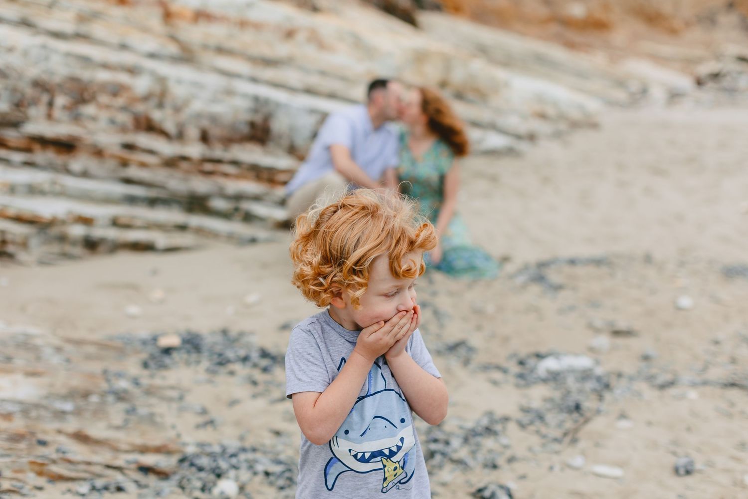 A young child in a gray t-shirt covers their mouth while standing on rocky coastal terrain with two people in background.