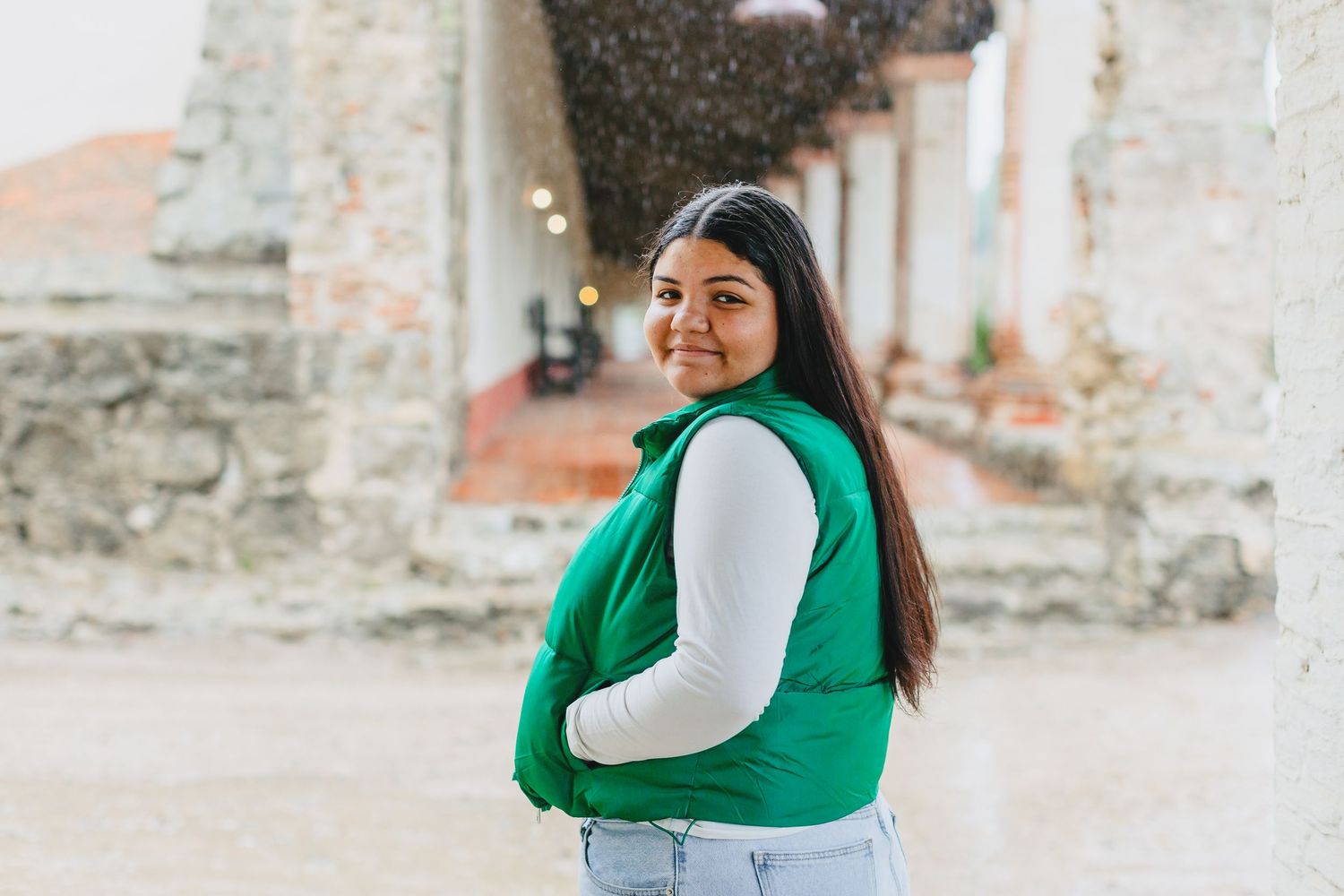 A person wearing a green vest and white long sleeve shirt poses casually against a weathered concrete wall.