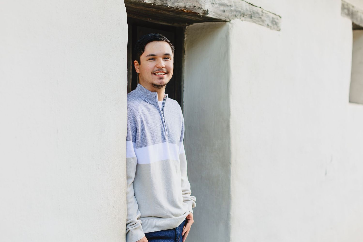 Someone in a light colored pullover with color blocking stands smiling in a white doorway.