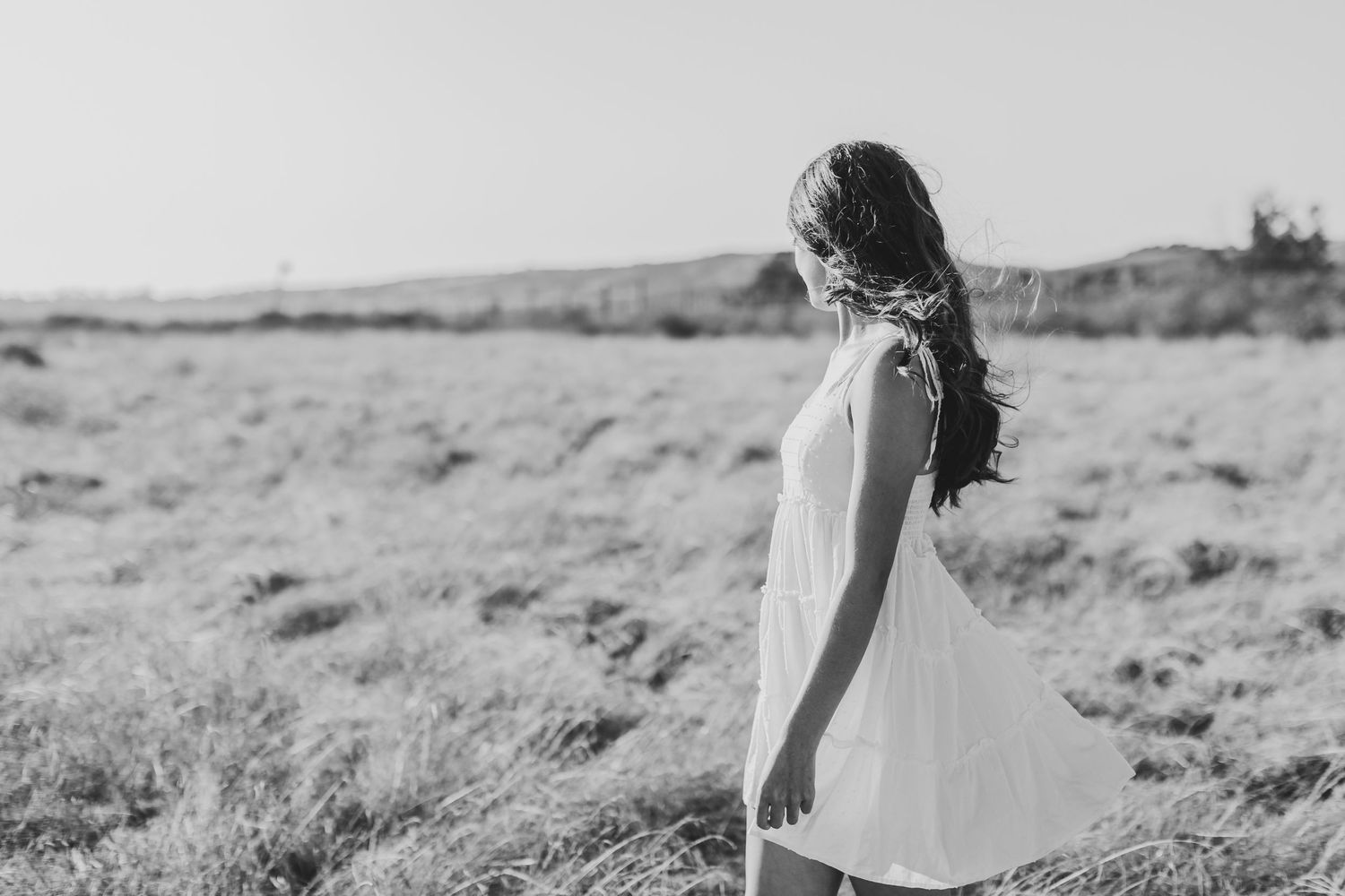 Person in white dress stands in profile against rolling hills landscape.