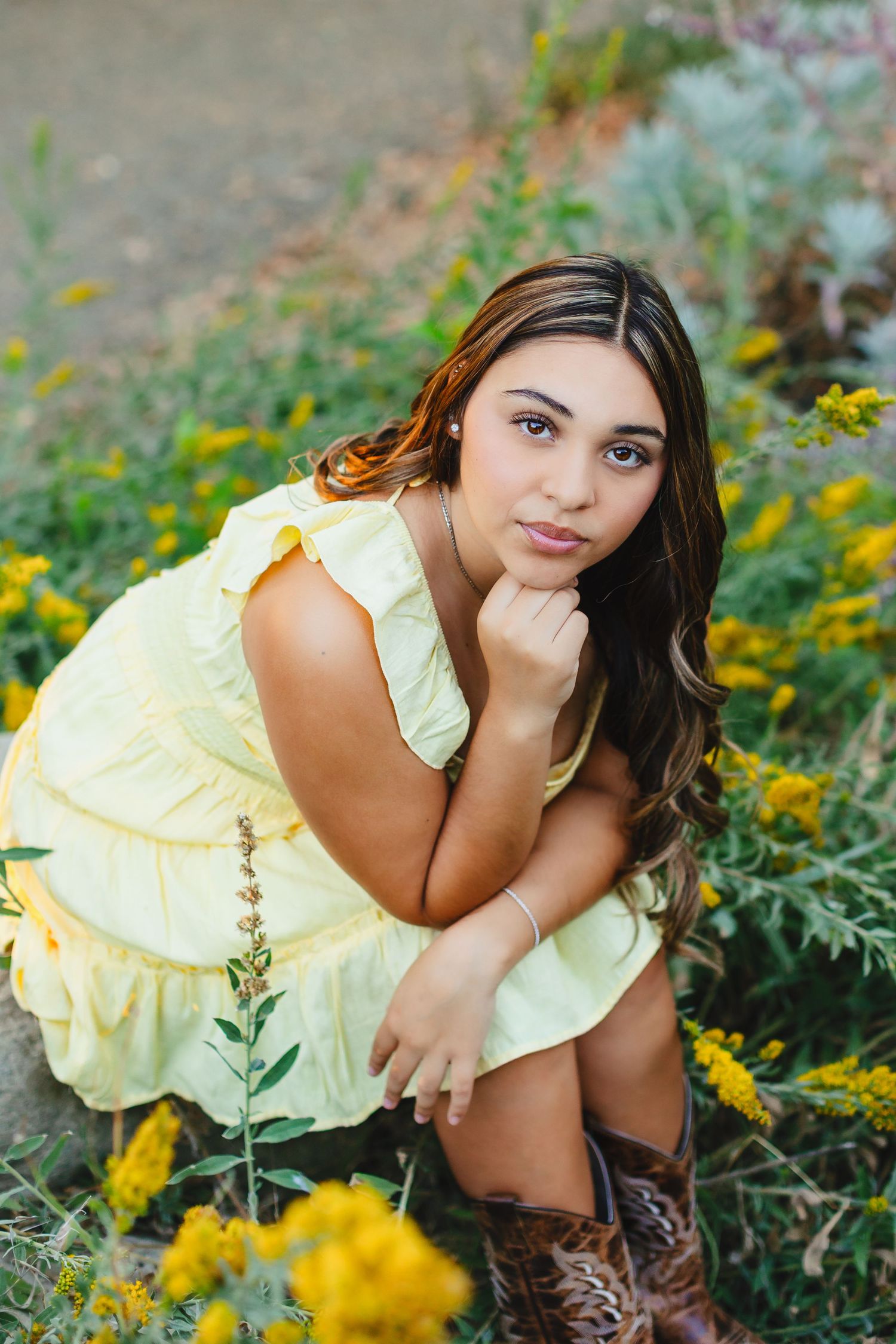 Young woman in yellow ruffle dress and cowboy boots crouching among yellow wildflowers in a natural outdoor setting.