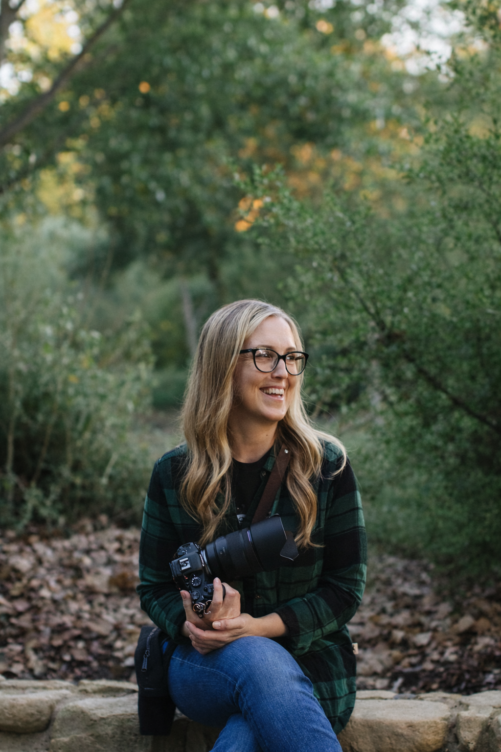 Smiling blonde woman with glasses holding a DSLR camera with telephoto lens, sitting outdoors in a lush green park.