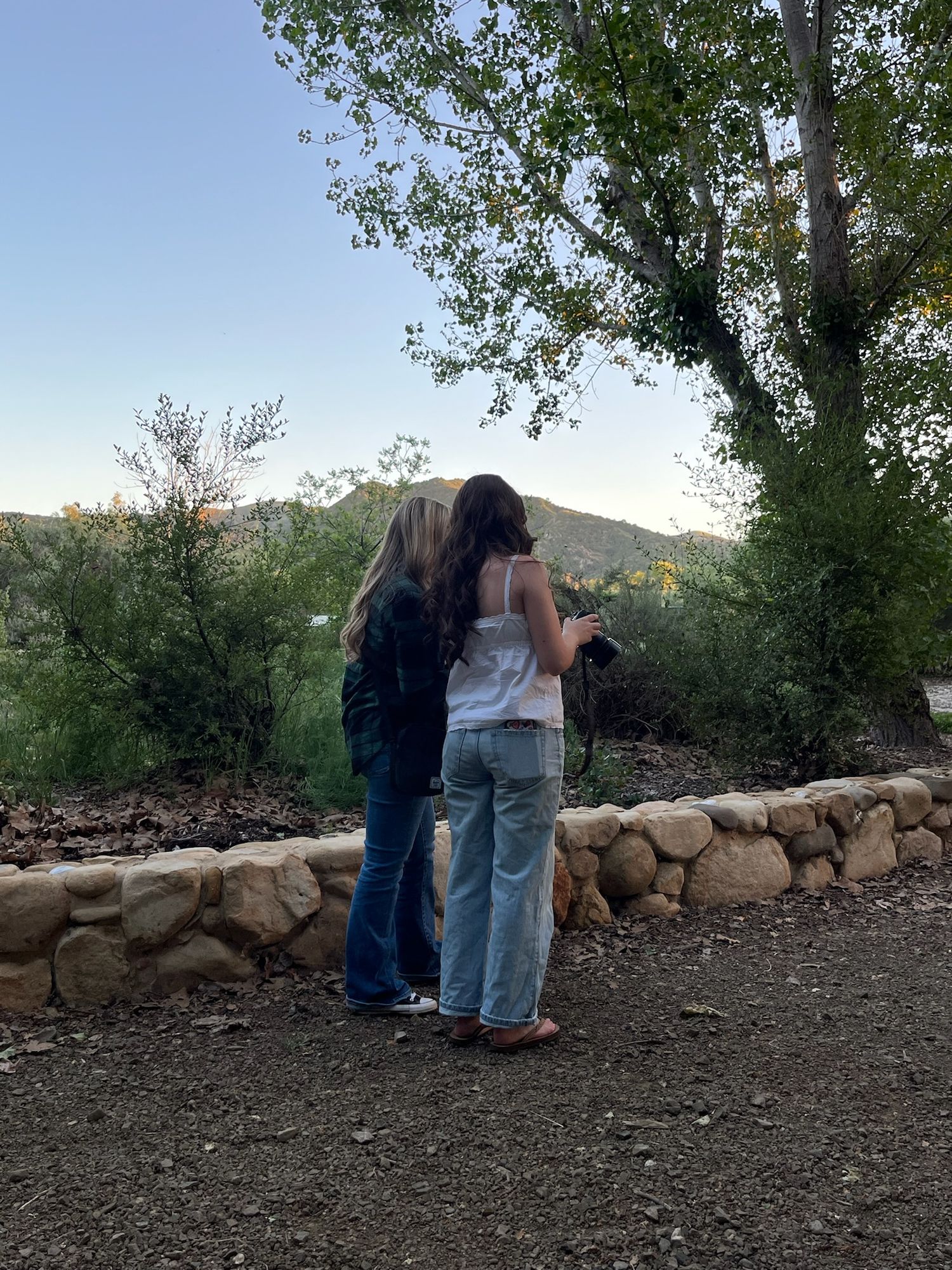 Two women stand together on a gravel path, looking at a camera near a stone wall with lush green hills in the background.