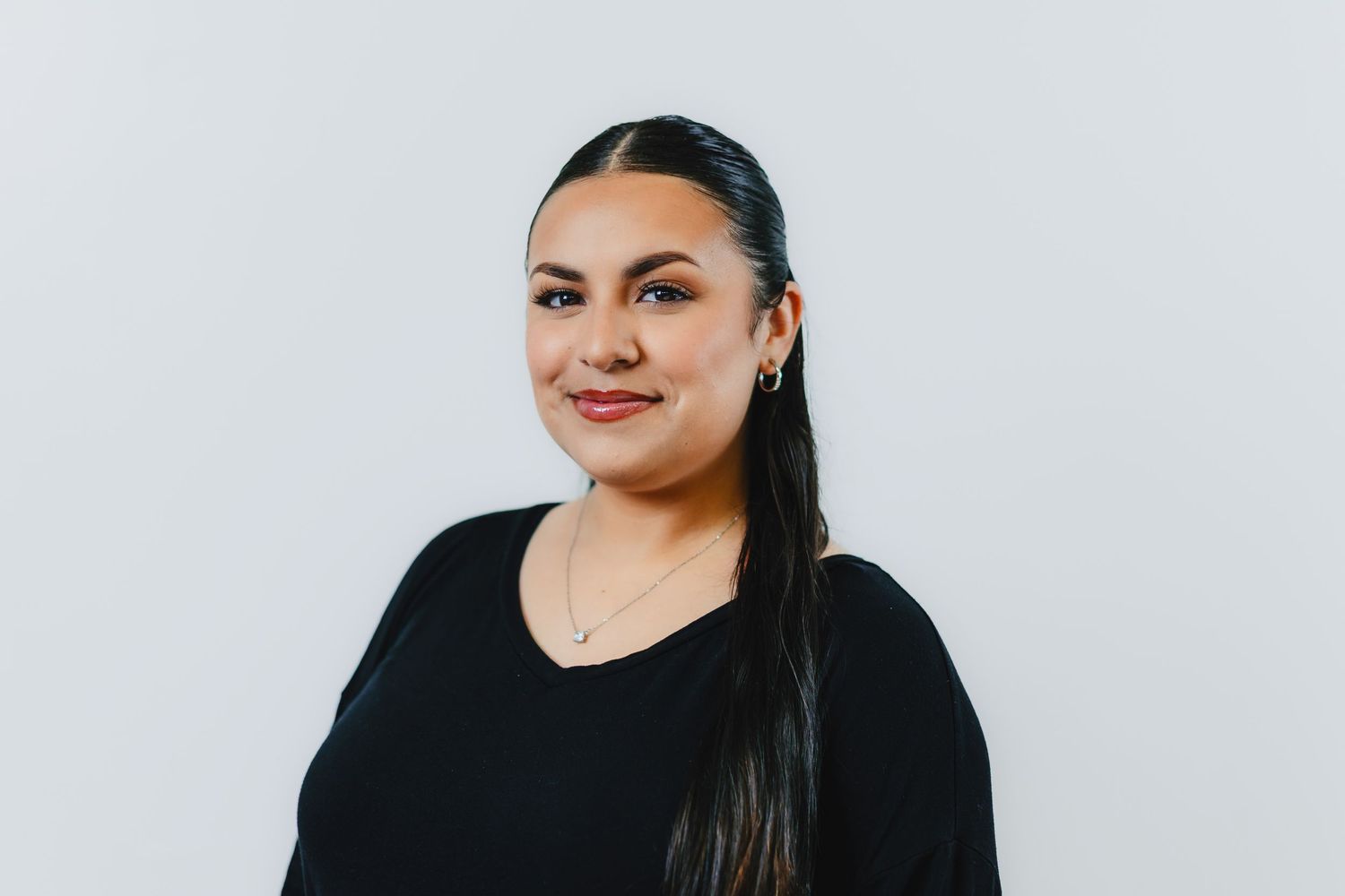 Professional headshot of someone wearing a black top against a white background, with long dark hair and a warm smile.