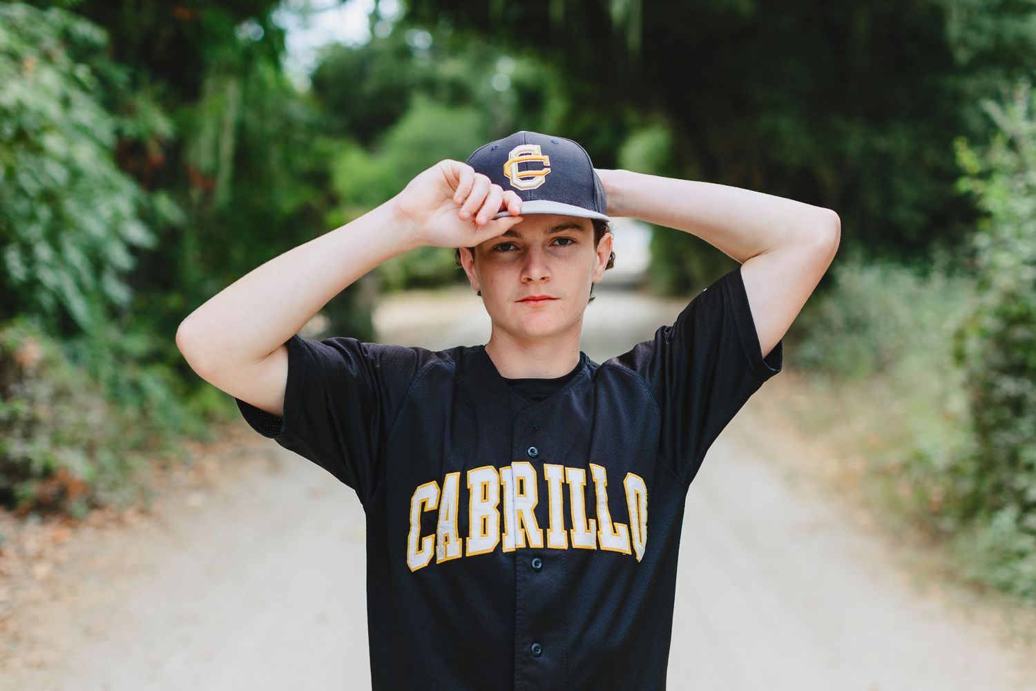 Teen boy in Cabrillo baseball jersey and cap posing on tree-lined dirt path outdoors.