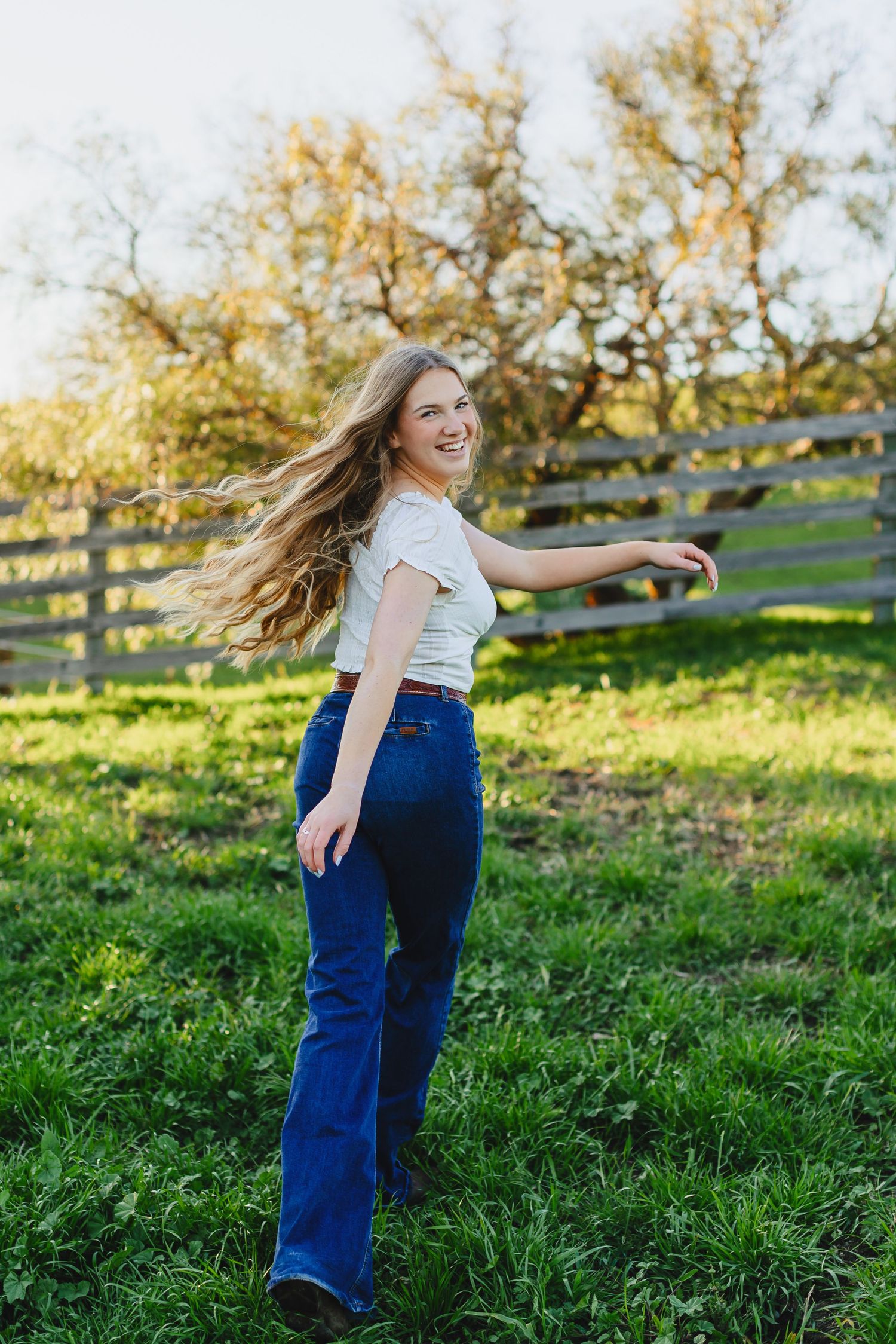 Woman in jeans and white top twirls happily in sunny field with wooden fence.