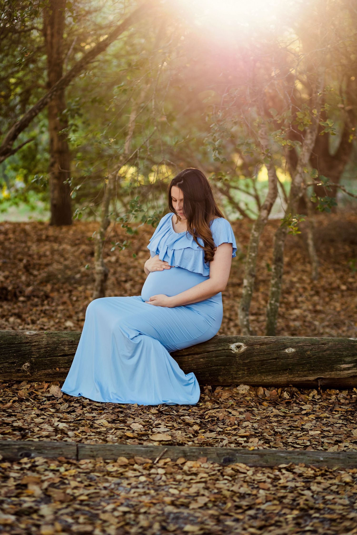 Person in blue dress sits contemplatively on fallen log surrounded by autumn leaves in sunlit wooded setting.