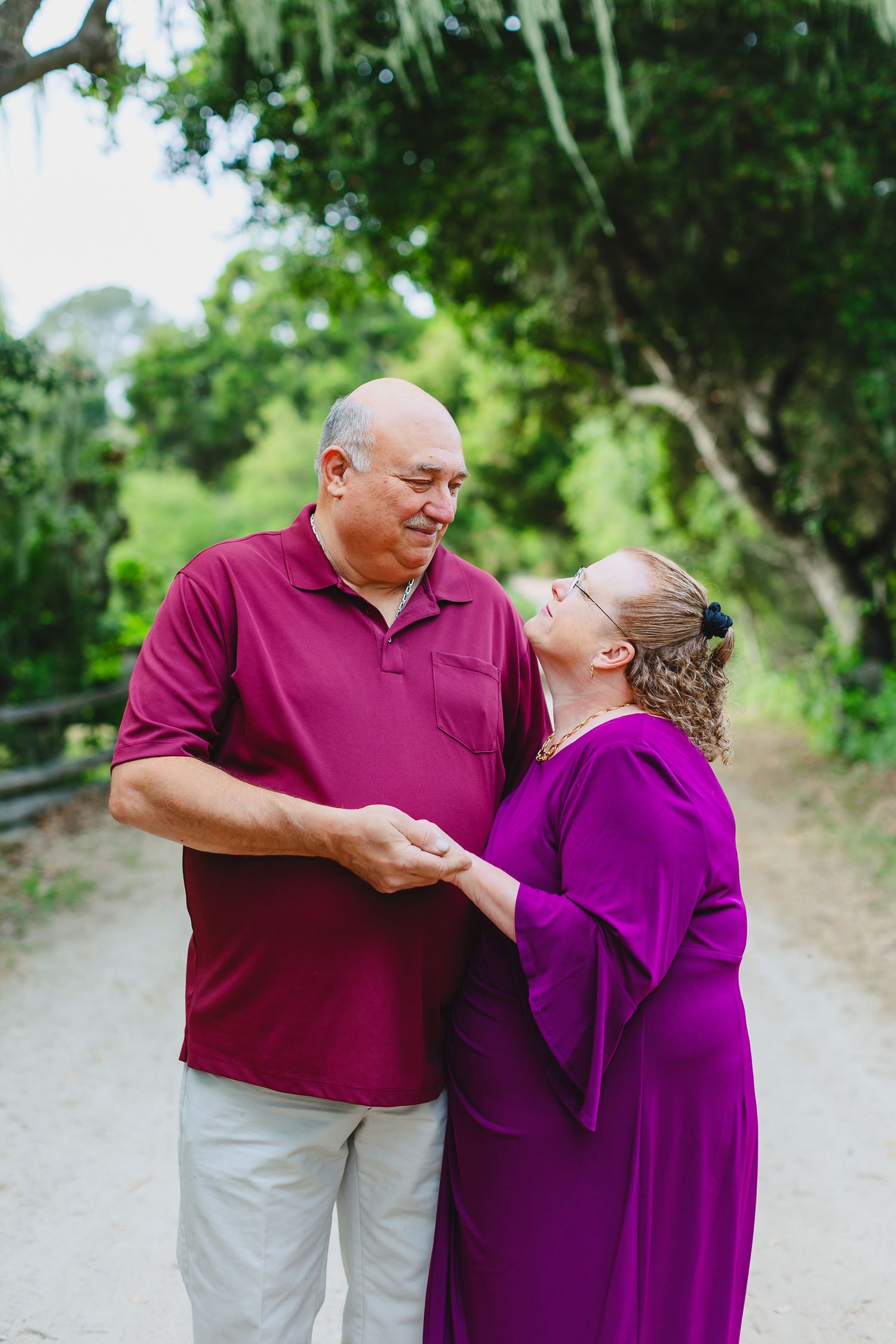 A couple in coordinating burgundy and purple outfits shares a tender moment while walking on a path surrounded by greenery.