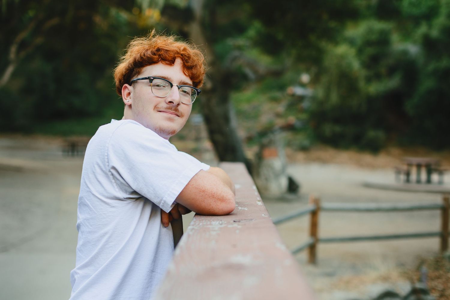 A person in glasses and white t-shirt leans against a wooden railing on an outdoor path surrounded by greenery.