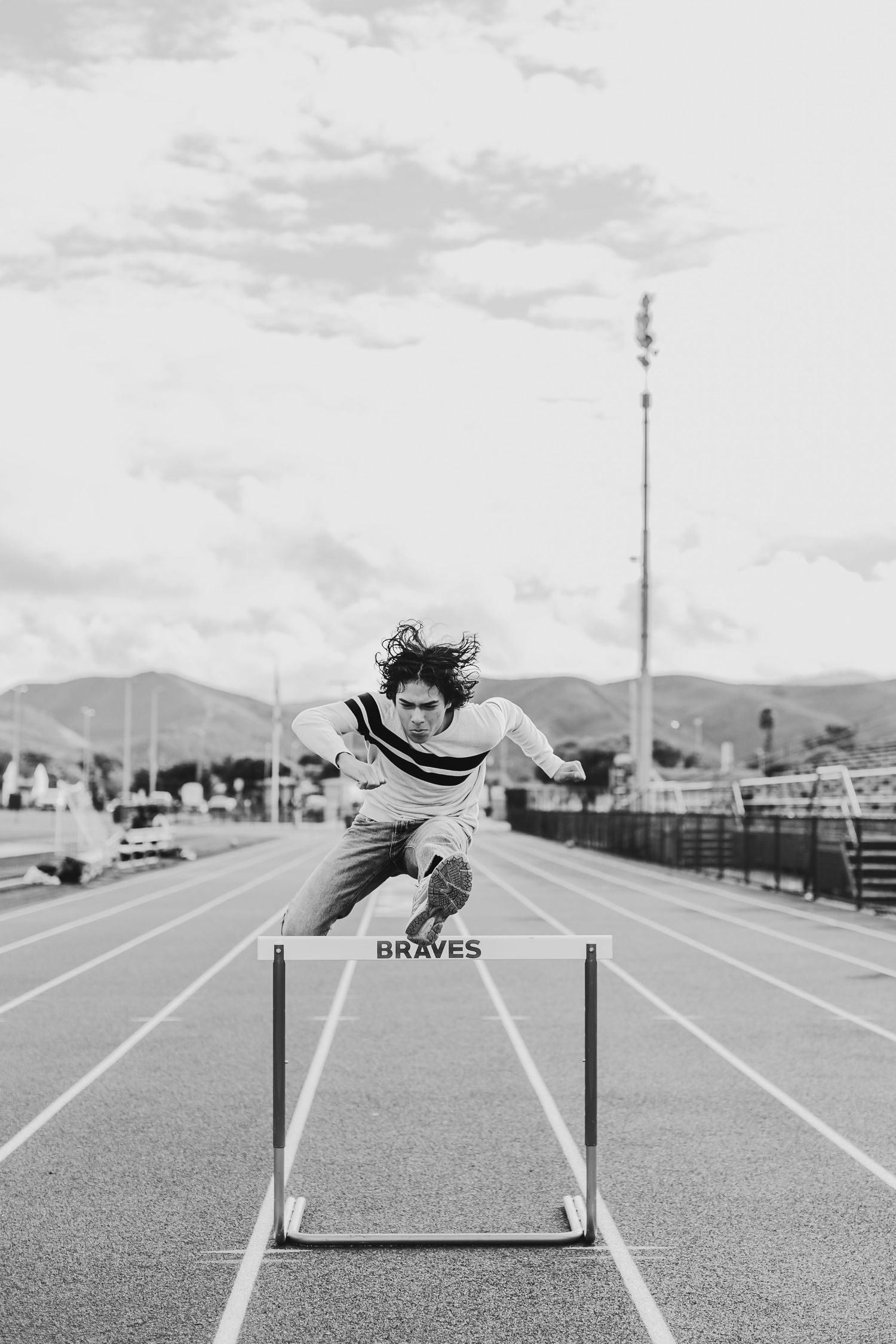 Black and white photo of an athlete jumping over a Braves hurdle on an outdoor track with mountains in the background.