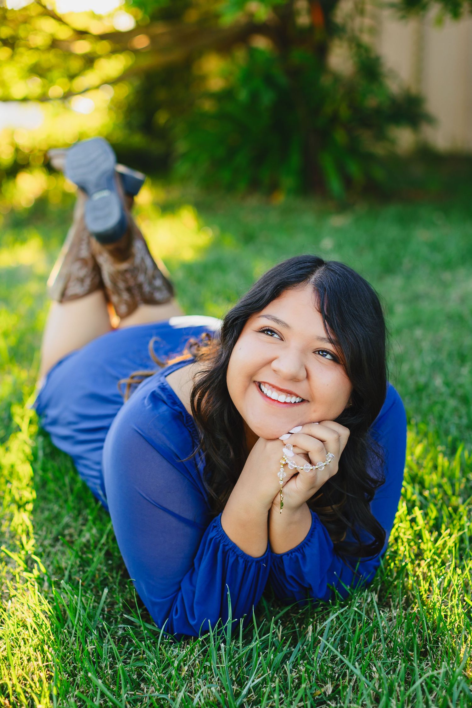 Young woman in blue dress lying on green grass, smiling brightly with hands clasped under chin on sunny day.
