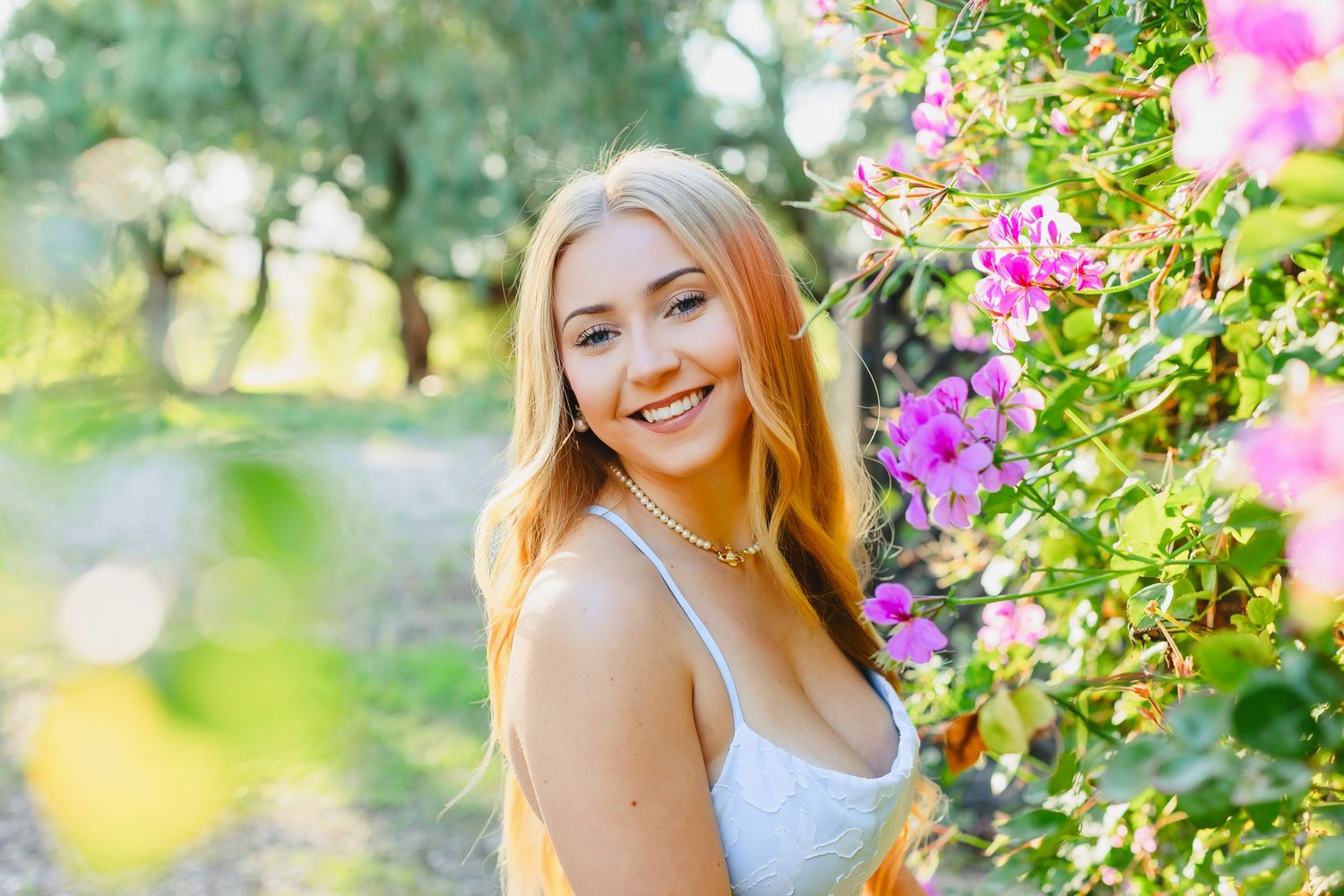 A person in a white top poses outdoors among vibrant pink flowers and green foliage in a sunny garden setting.