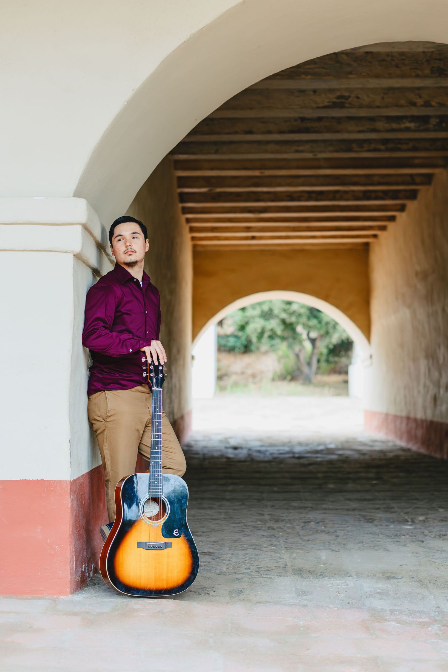 Young man in burgundy shirt leans against an arched adobe corridor wall holding an acoustic guitar.