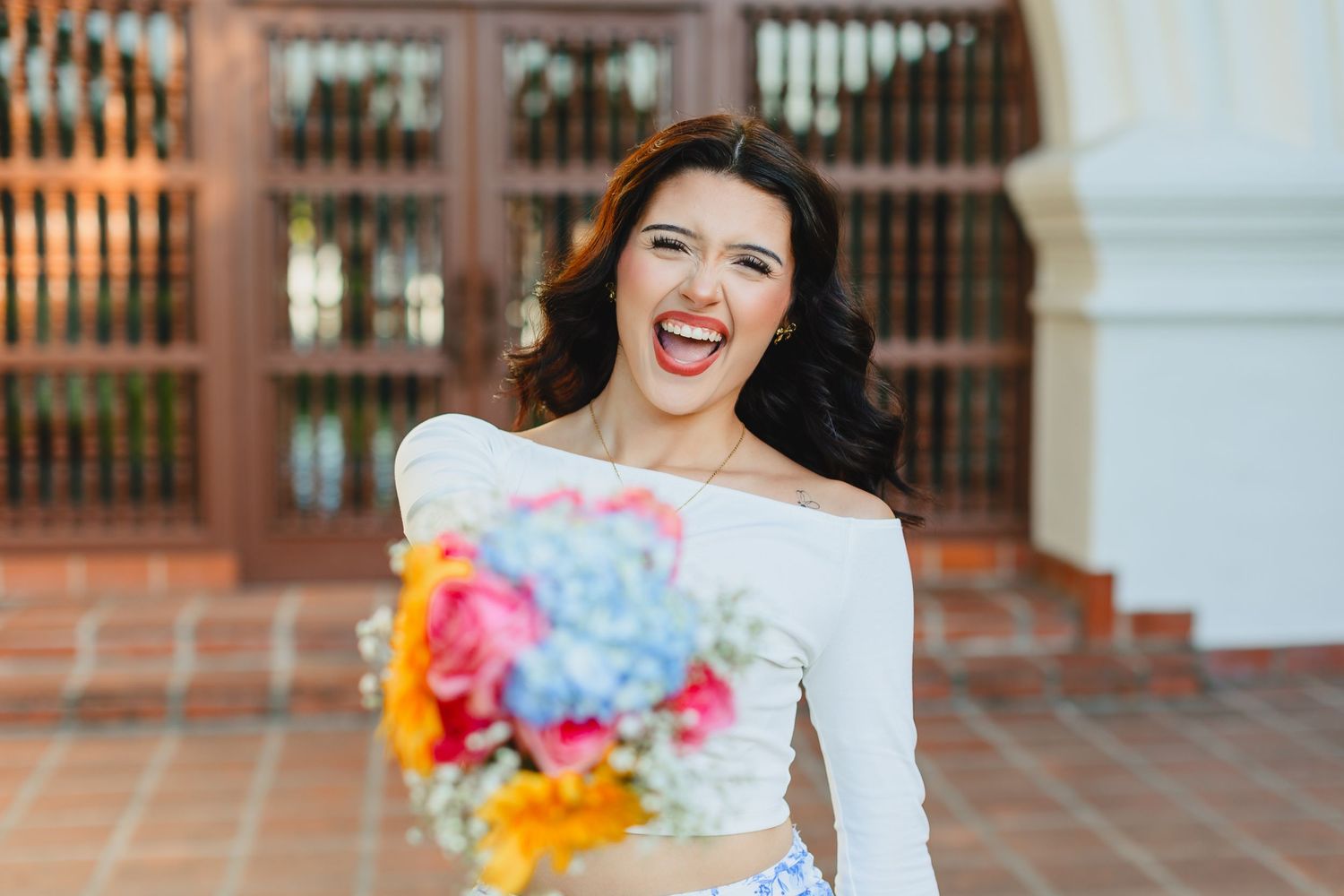 A joyful laugh while holding a vibrant pink, orange and blue bouquet and wearing a white off-shoulder dress.